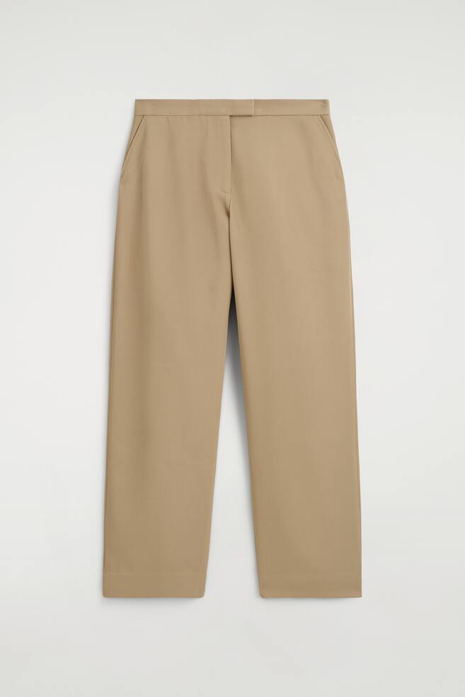 Beige Hosen sind von vorne zu sehen, mit einem sauberen Bund, einem verdeckten Frontverschluss und seitlichen Eingrifftaschen. Die Hosen präsentieren einen entspannten, geraden Schnitt mit einer Bügelfalte entlang jedes Beins. - 1