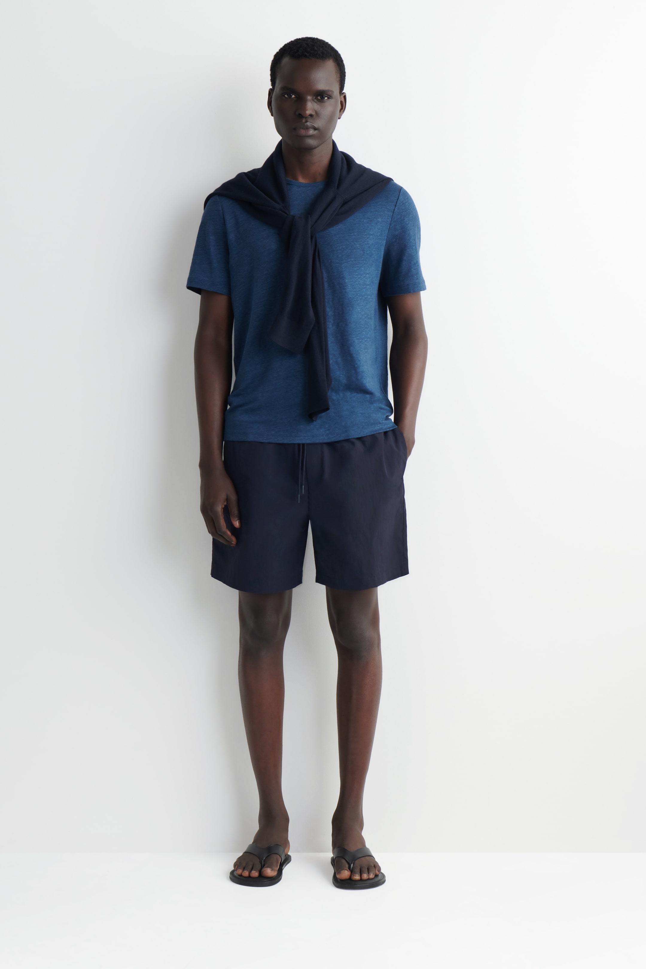 Visa större bild: En blåmelerad t-shirt med rund hals bärs, med en marinblå tröja draperad över axlarna och knuten framtill. Den kombineras med marinblå shorts med synlig dragsko i midjan och svarta flip-flops i remstil.