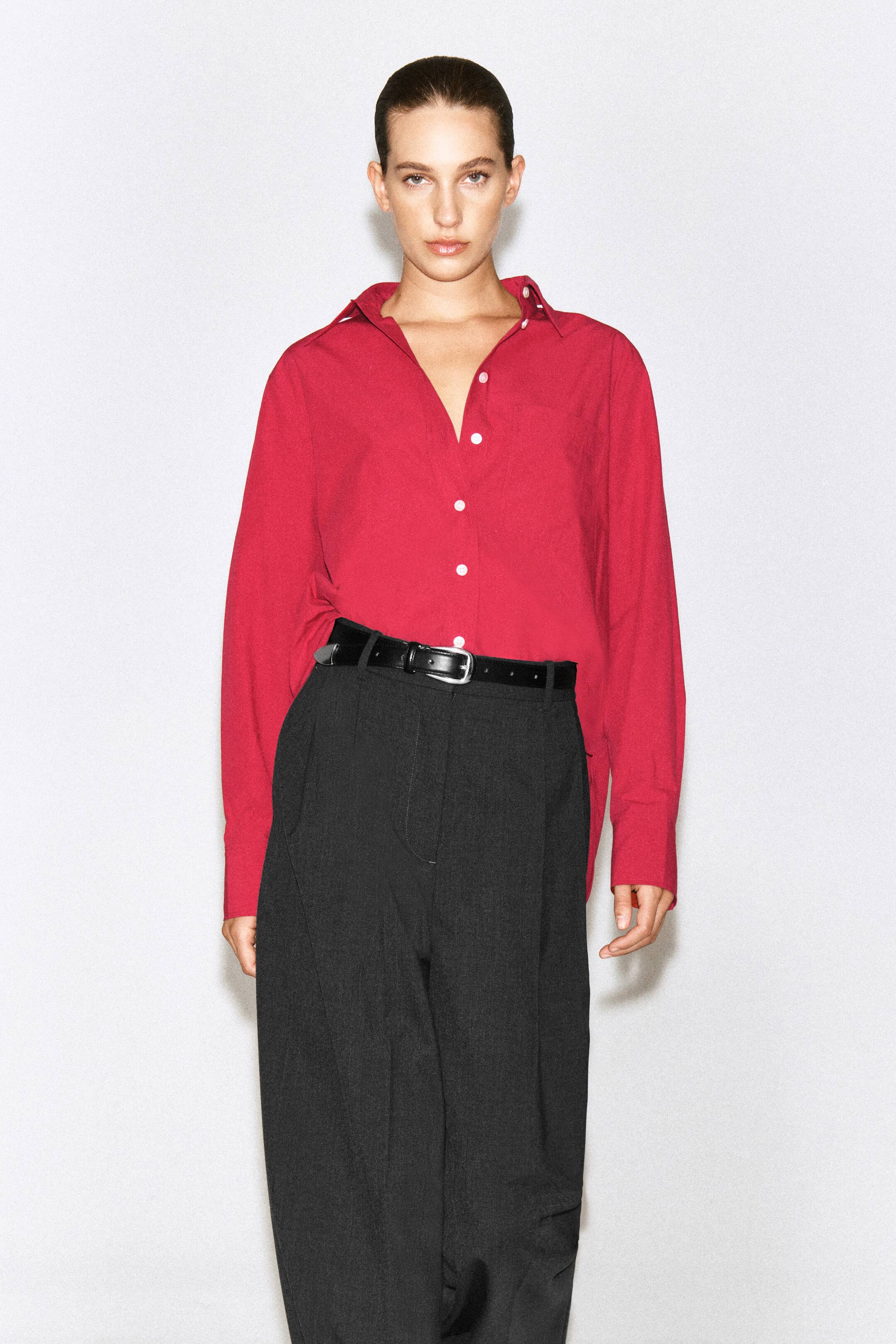Agrandir l'image: Une chemise rouge à manches longues et boutonnée est portée par le mannequin, rentrée dans un pantalon foncé avec une ceinture noire. La chemise présente des boutons blancs sur le devant, un col pointu et se porte avec les premiers boutons laissés ouverts.