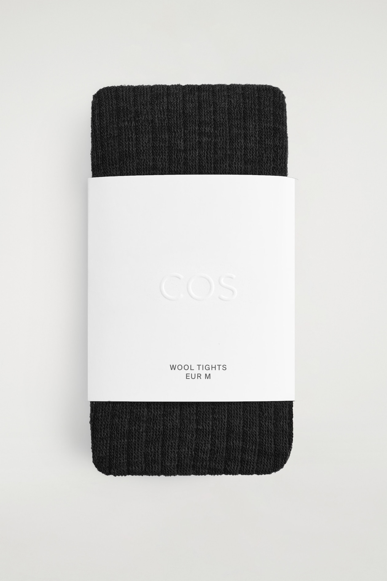 A pair of folded, dark charcoal wool tights is facing forward, displaying a distinct ribbed knit texture. The tights are wrapped centrally in a wide white paper band, showcasing an embossed "COS" logo and printed text reading "WOOL TIGHTS EUR M."