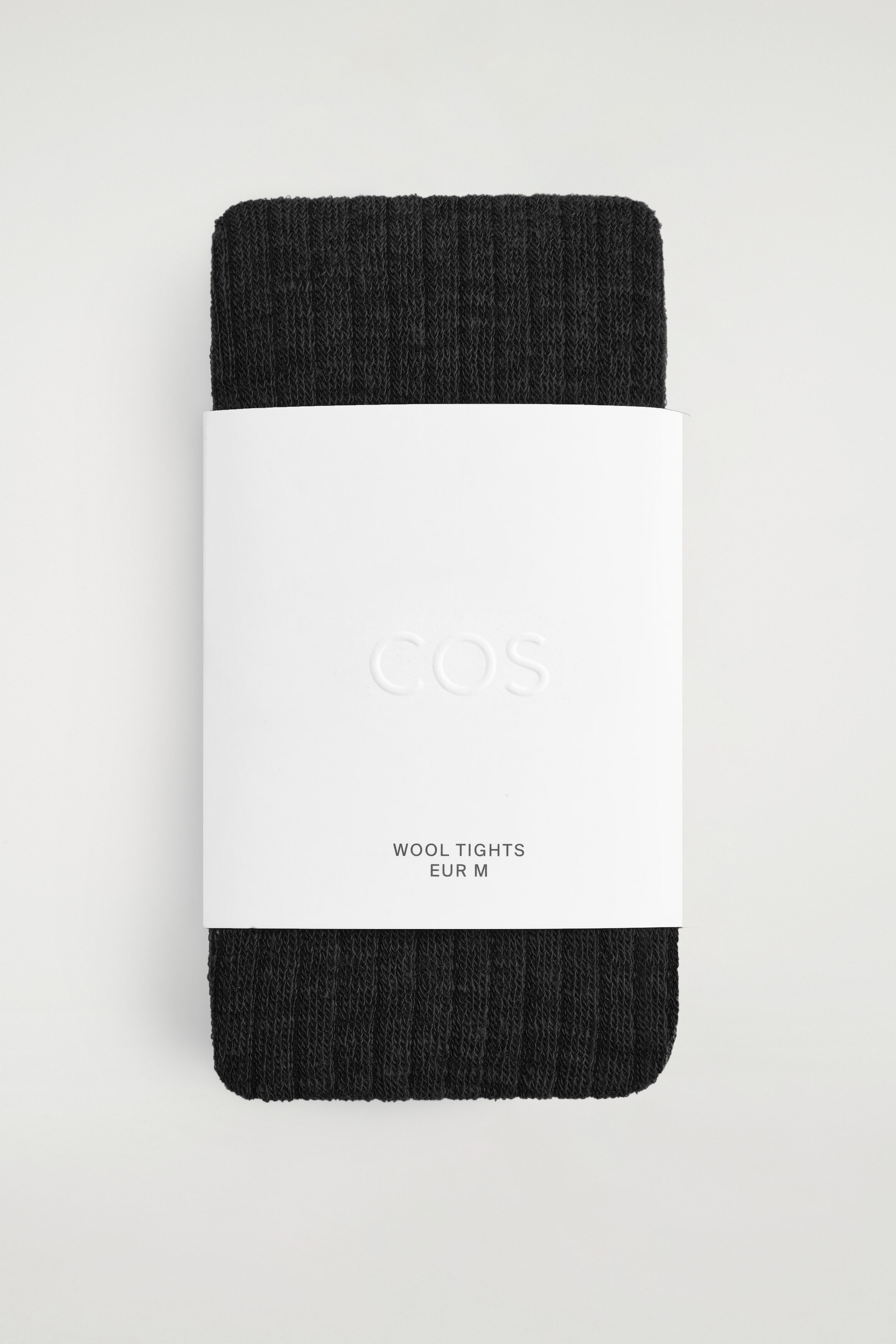 A pair of folded, dark charcoal wool tights is facing forward, displaying a distinct ribbed knit texture. The tights are wrapped centrally in a wide white paper band, showcasing an embossed "COS" logo and printed text reading "WOOL TIGHTS EUR M."