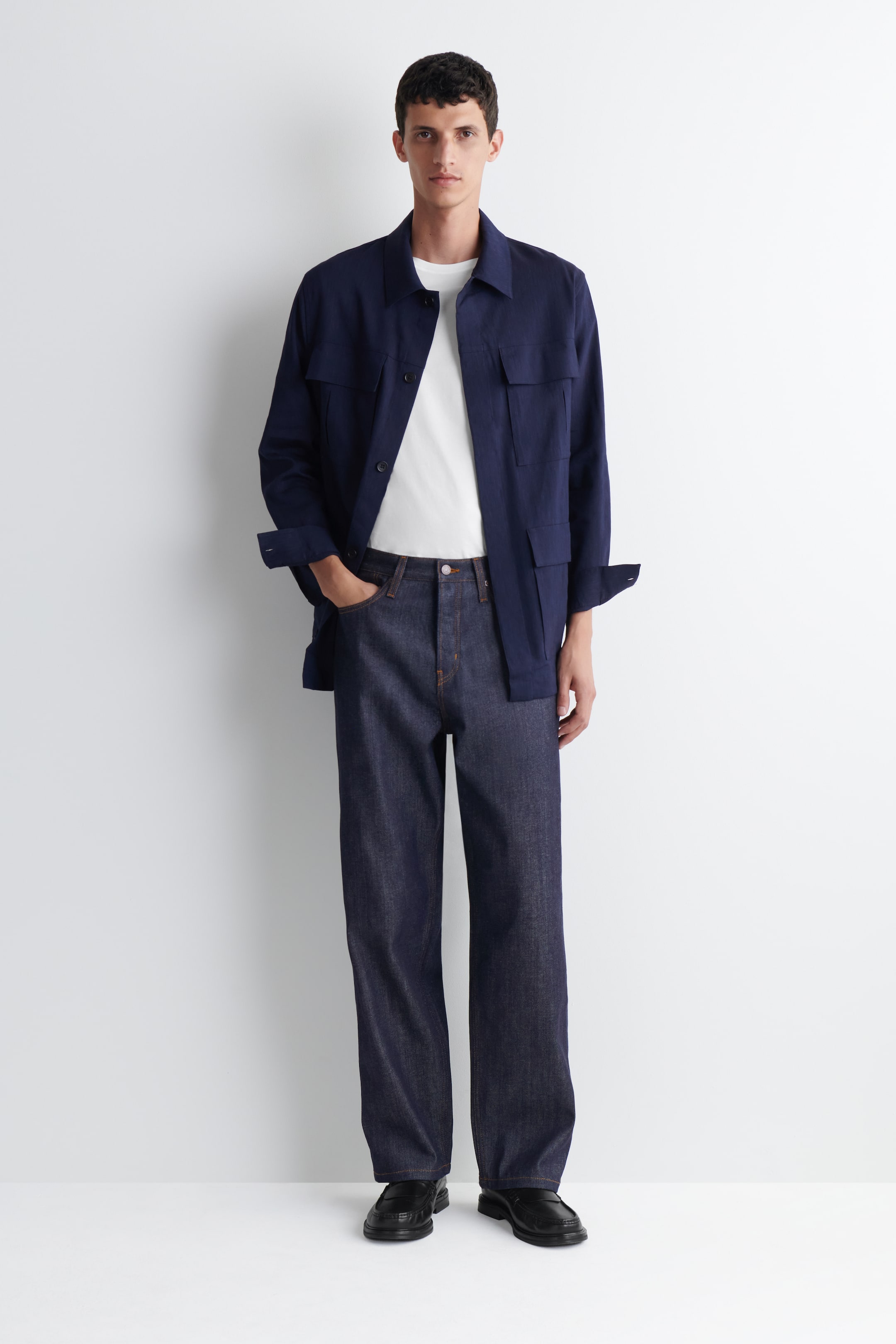 Grotere afbeelding bekijken: Een marineblauwe overshirt met een klassieke kraag en twee borstzakken en twee lagere klepzakken. De lange mouwen zijn opgerold en het wordt open gedragen over een wit T-shirt met ronde hals.