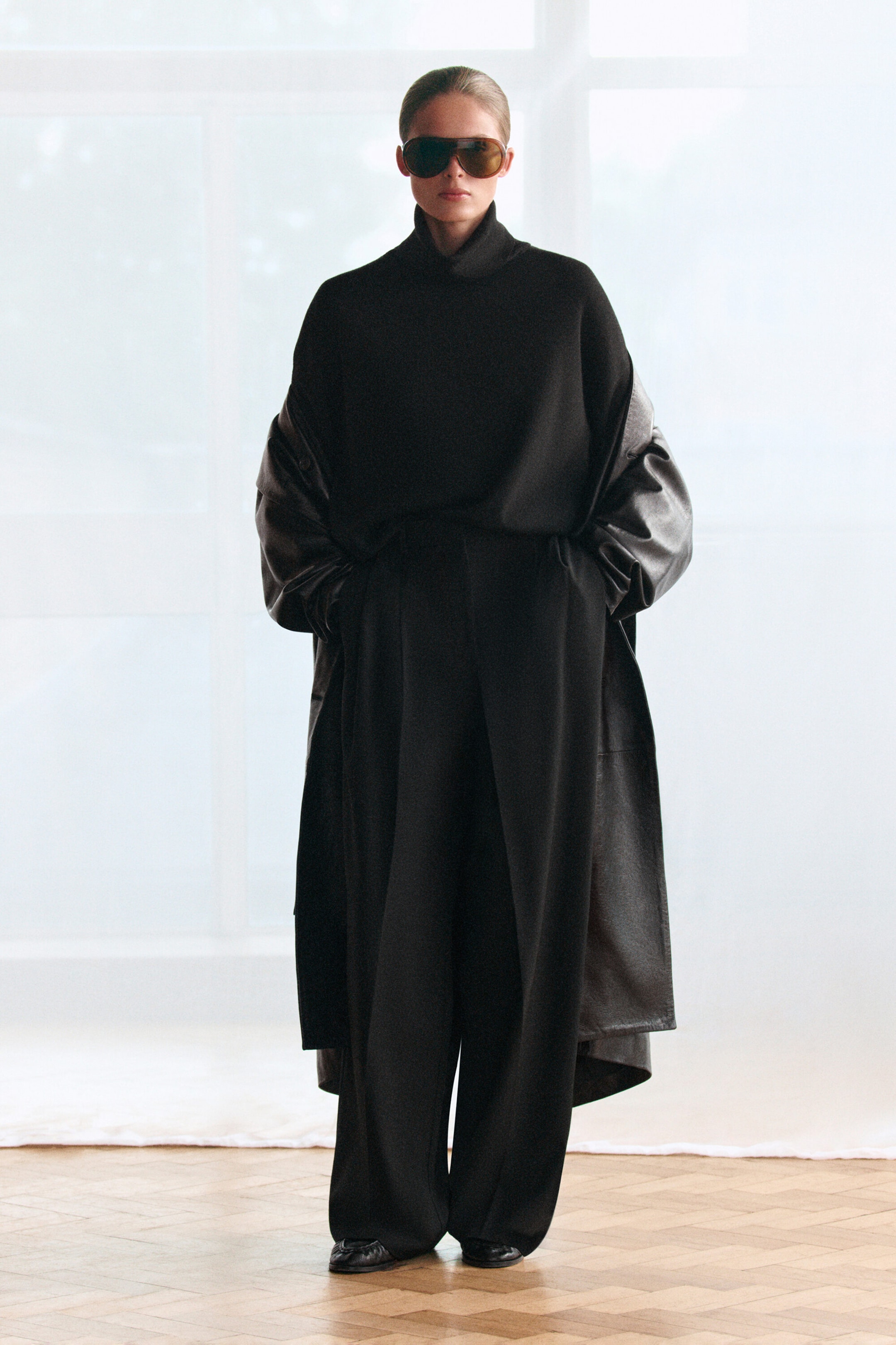 Der schwarze Rollkragenpullover aus Merinowolle ist von vorne zu sehen und zeichnet sich durch eine oversized, lässige Silhouette sowie einen hohen, strukturierten Rollkragen aus. Der Pullover ist in eine weite Hose gesteckt und unter einem drapierten Ledercoat geschichtet, wodurch sein weicher Fall und die feine Strickstruktur betont werden.