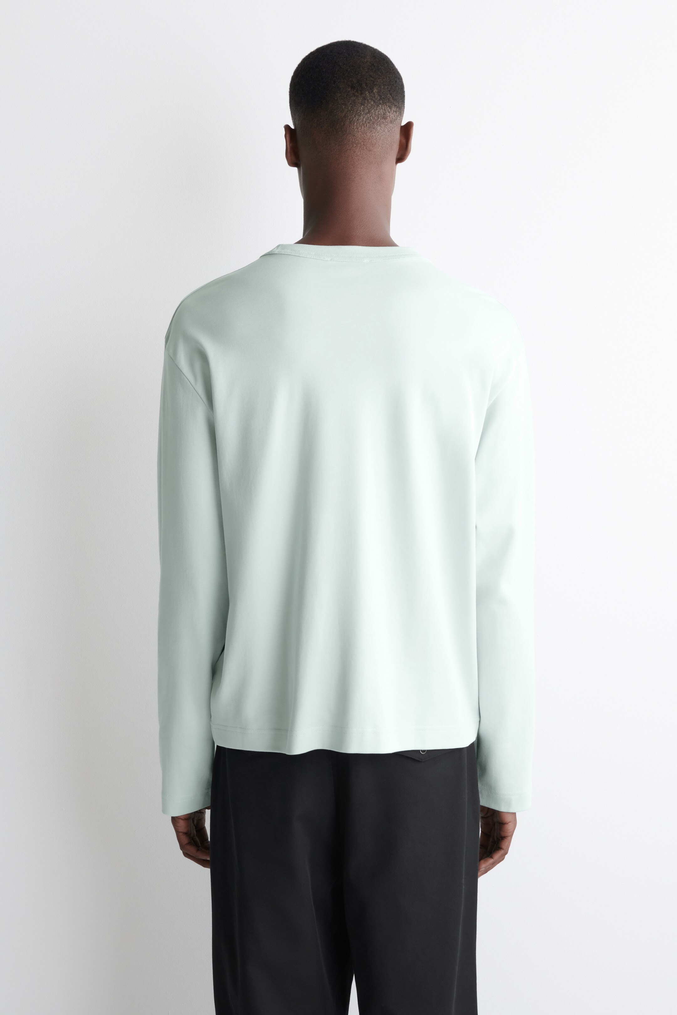 View larger image: A light green long-sleeved shirt is being presented from the back. It is displaying a crew neck, long sleeves, and a relaxed fit with a straight hem, and is being worn over black trousers.
