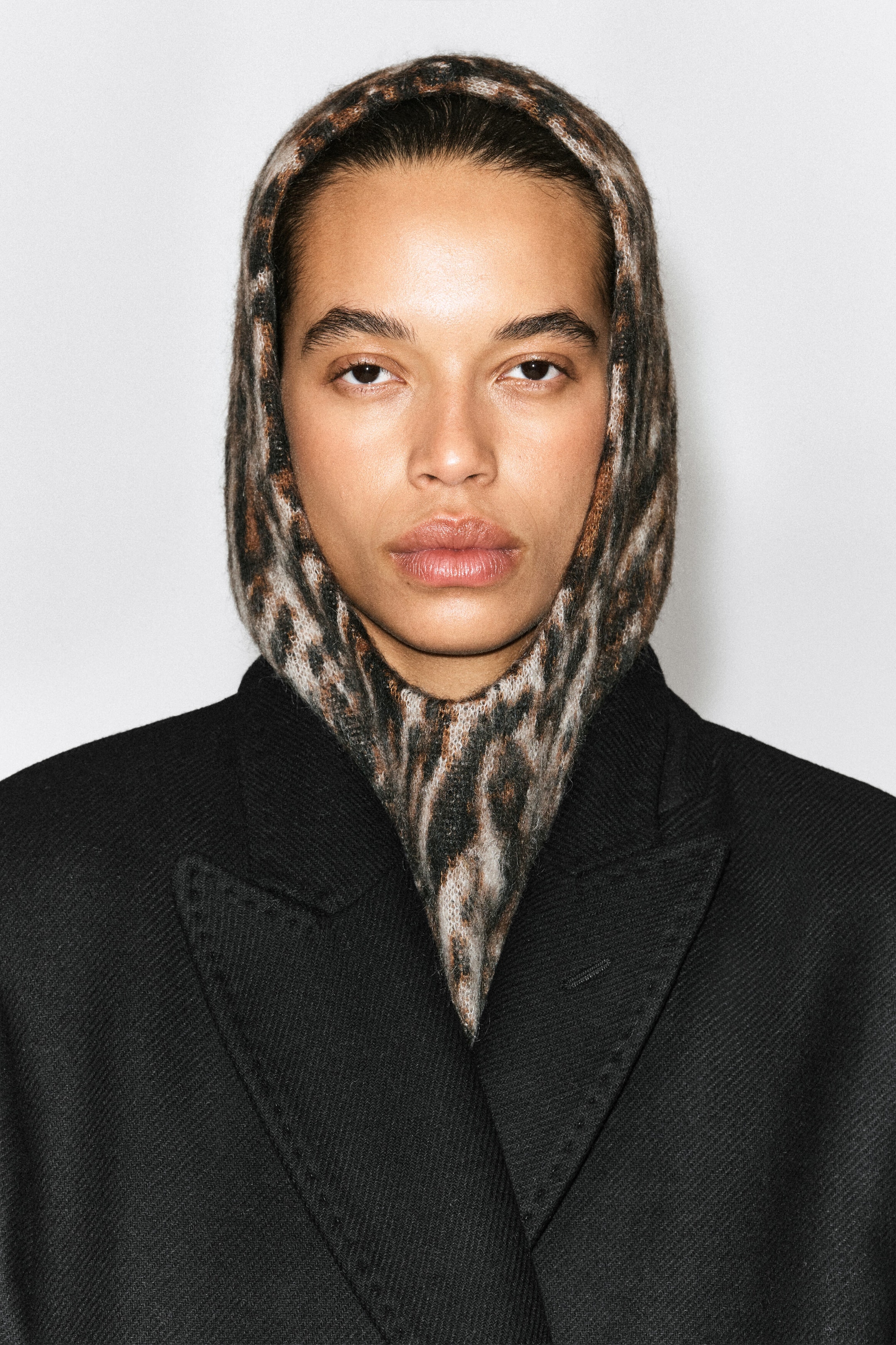 The leopard-jacquard balaclava is shown from the front, closely framing the face and covering the neck with its soft, fuzzy knit fabric. It features a brown and black leopard print pattern and is worn layered underneath a black coat with structured lapels. The leopard-jacquard balaclava is shown from the front, closely framing the face and covering the neck with its soft, fuzzy knit fabric. It features a brown and black leopard print pattern and is worn layered underneath a black coat with structured lapels.