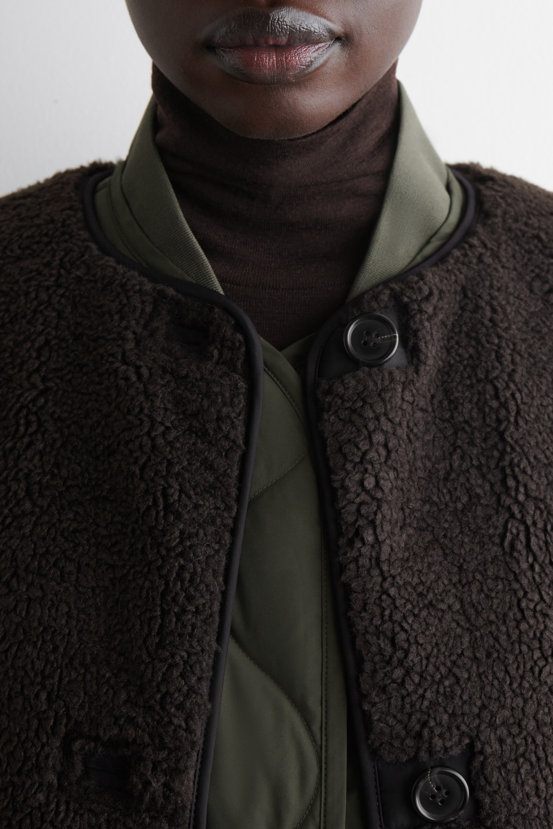 Agrandir l'image: Une veste en sherpa texturée de couleur brun foncé est ornée d'un passepoil noir le long de son encolure arrondie et de son ouverture frontale, où un bouton noir est visible. Elle est portée ouverte sur une veste matelassée vert olive, révélant son col côtelé et sa texture matelassée.
