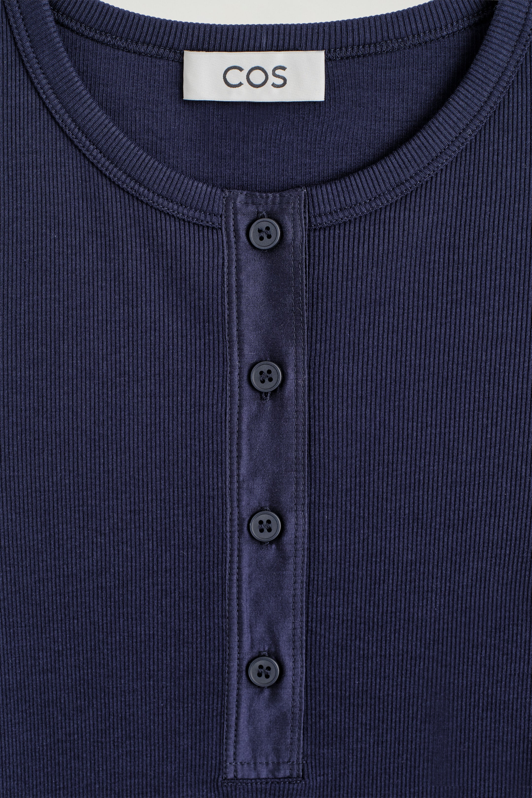 This close-up front view of the navy blue ribbed cotton Henley top shows a round neckline with a ribbed collar and a central button placket. The placket features four matching dark buttons on a smooth fabric trim, contrasting with the garment’s fine, vertical ribbed knit texture. This close-up front view of the navy blue ribbed cotton Henley top shows a round neckline with a ribbed collar and a central button placket. The placket features four matching dark buttons on a smooth fabric trim, contrasting with the garment’s fine, vertical ribbed knit texture.