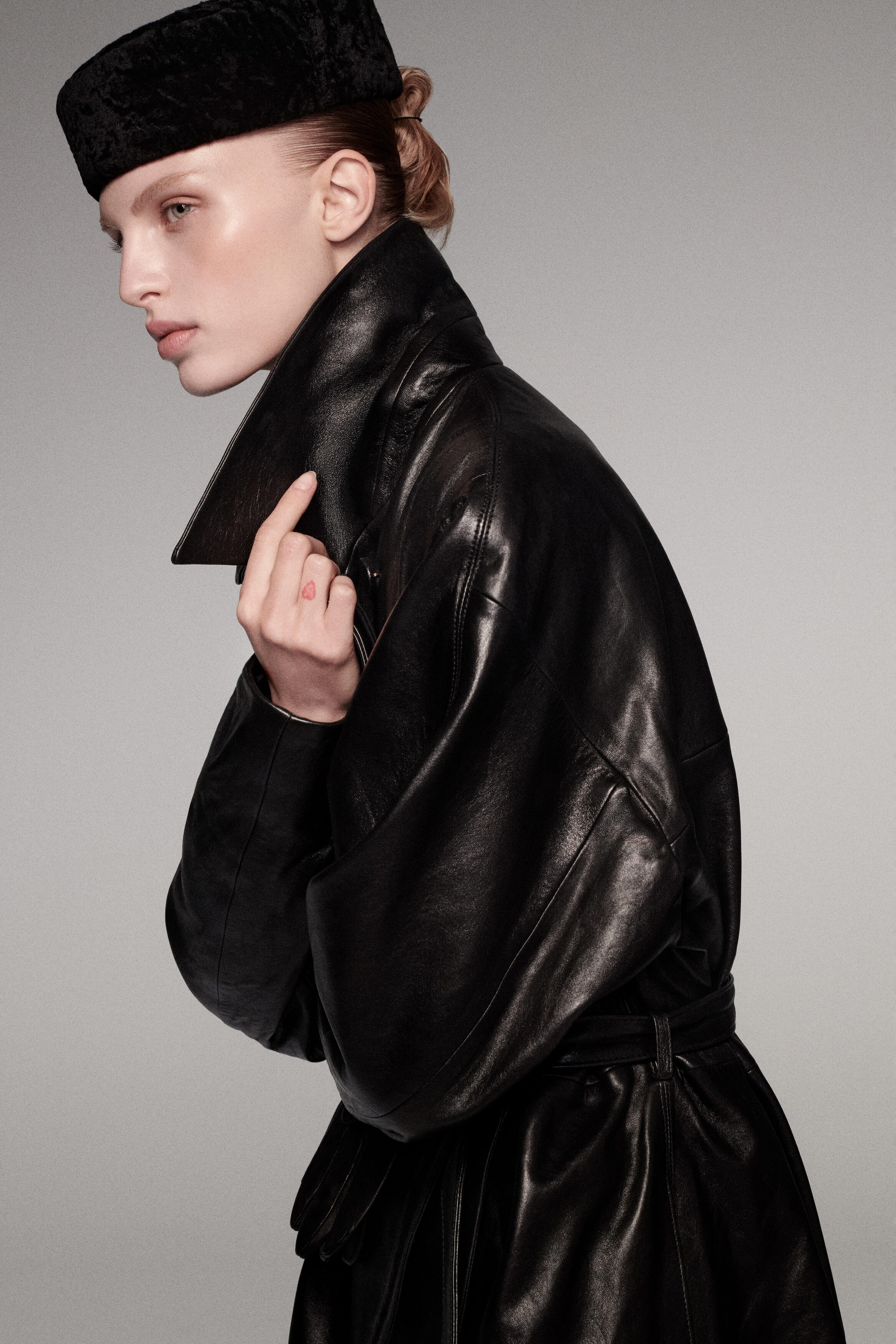 An oversized belted leather long coat is shown in side profile, highlighting its voluminous silhouette, dropped shoulders, and cinched belted waist. The high, structured collar is held upright, showcasing the smooth, semi-glossy texture of the black leather. An oversized belted leather long coat is shown in side profile, highlighting its voluminous silhouette, dropped shoulders, and cinched belted waist. The high, structured collar is held upright, showcasing the smooth, semi-glossy texture of the black leather.