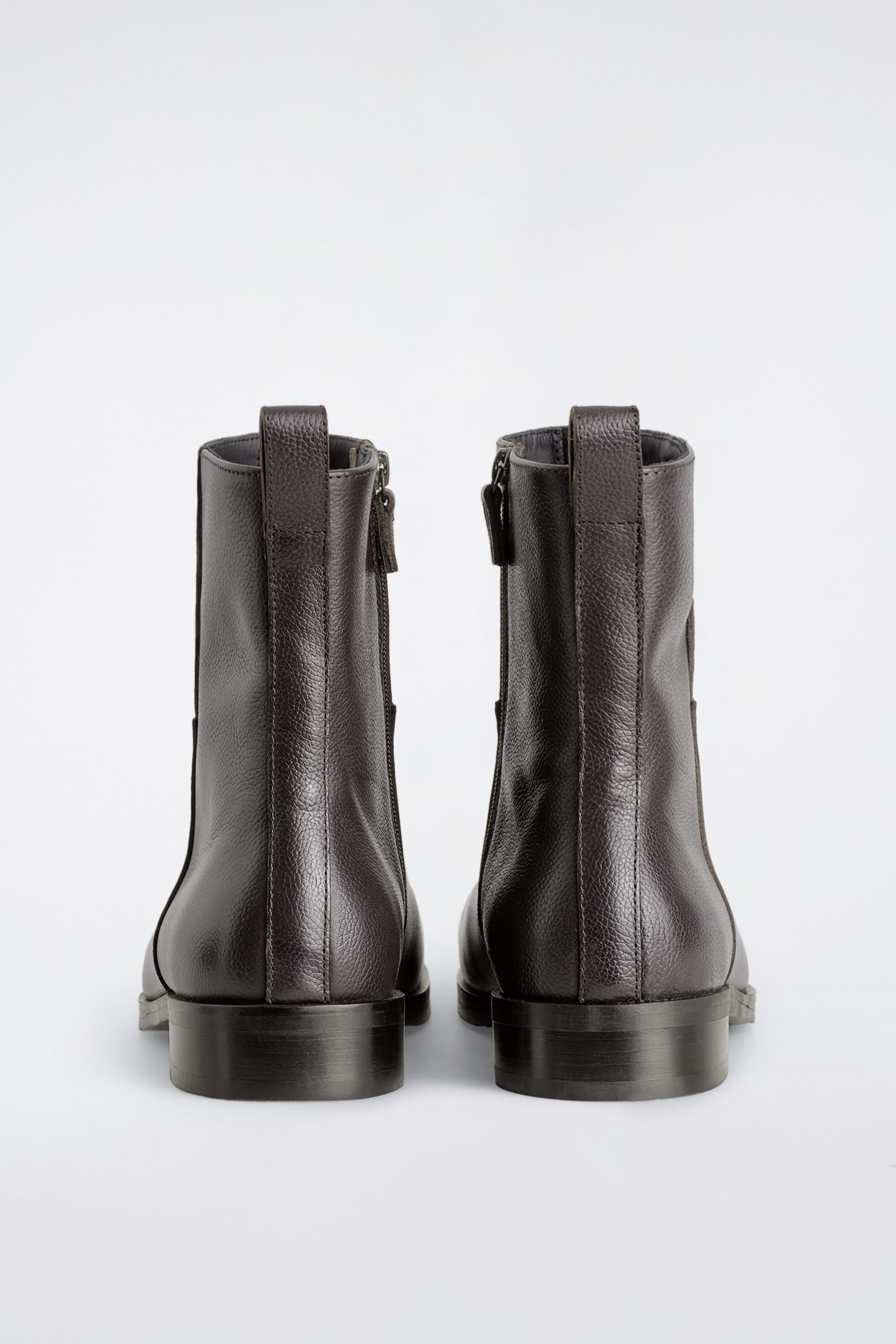 Square-toe leather ankle boots are shown from the back, displaying their dark brown grained leather texture, vertical rear seams with pull tabs, and low stacked block heels. Inner-side zip closures extend along the shafts of both boots.