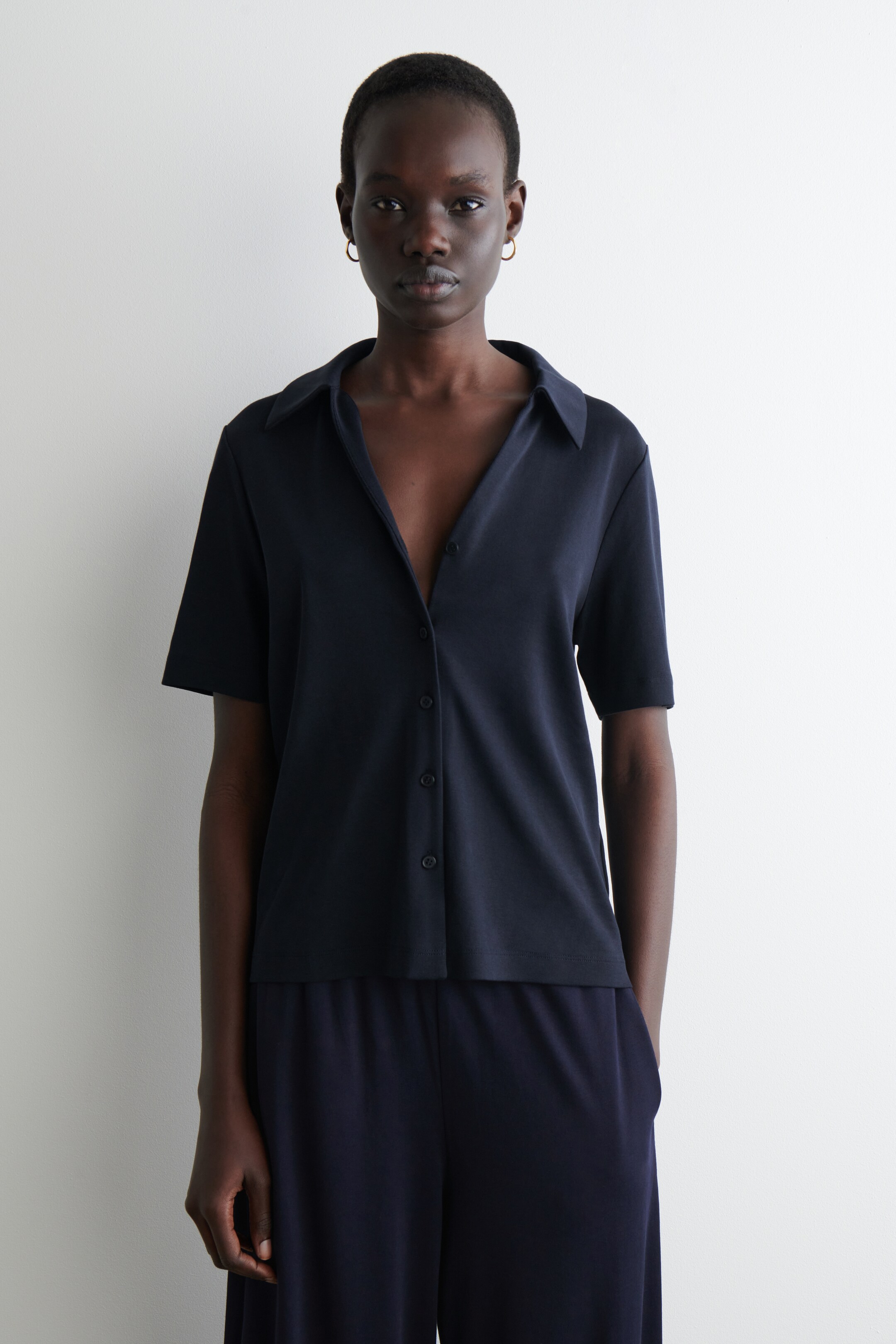 Agrandir l'image: Une chemise bleu marine à manches courtes en tissu à texture lisse est portée, avec un col pointu et une patte de boutonnage partiellement déboutonnée. Elle est assortie à un pantalon large bleu marine, formant un ensemble coordonné.