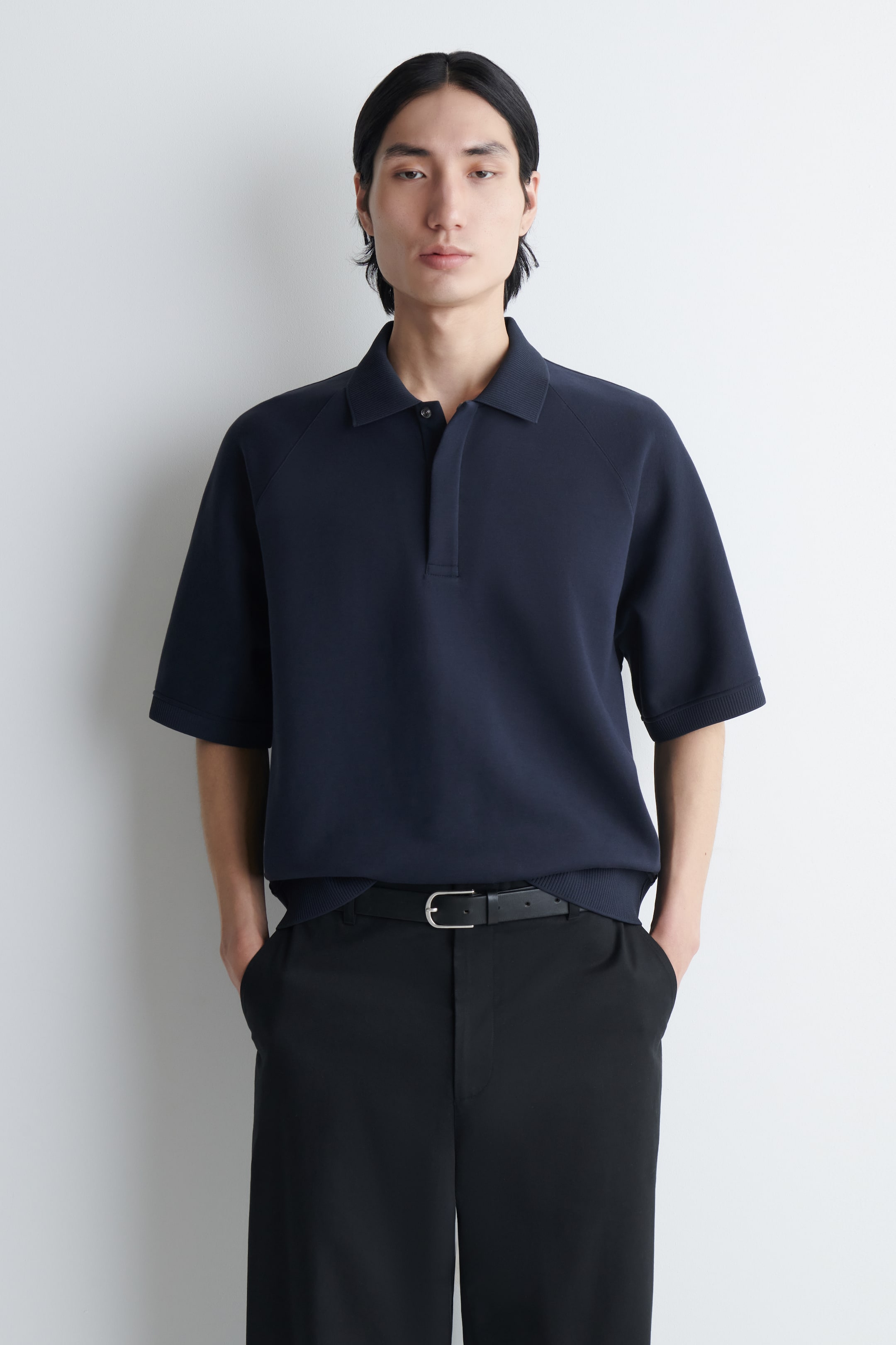 Vezi imaginea mai mare: Un tricou polo din jersey scuba, albastru marin, cu mâneci raglan scurte, guler clasic și fenta ascunsă, este purtat de model. Tricoul polo este introdus în pantaloni negri, asortați cu o curea neagră.