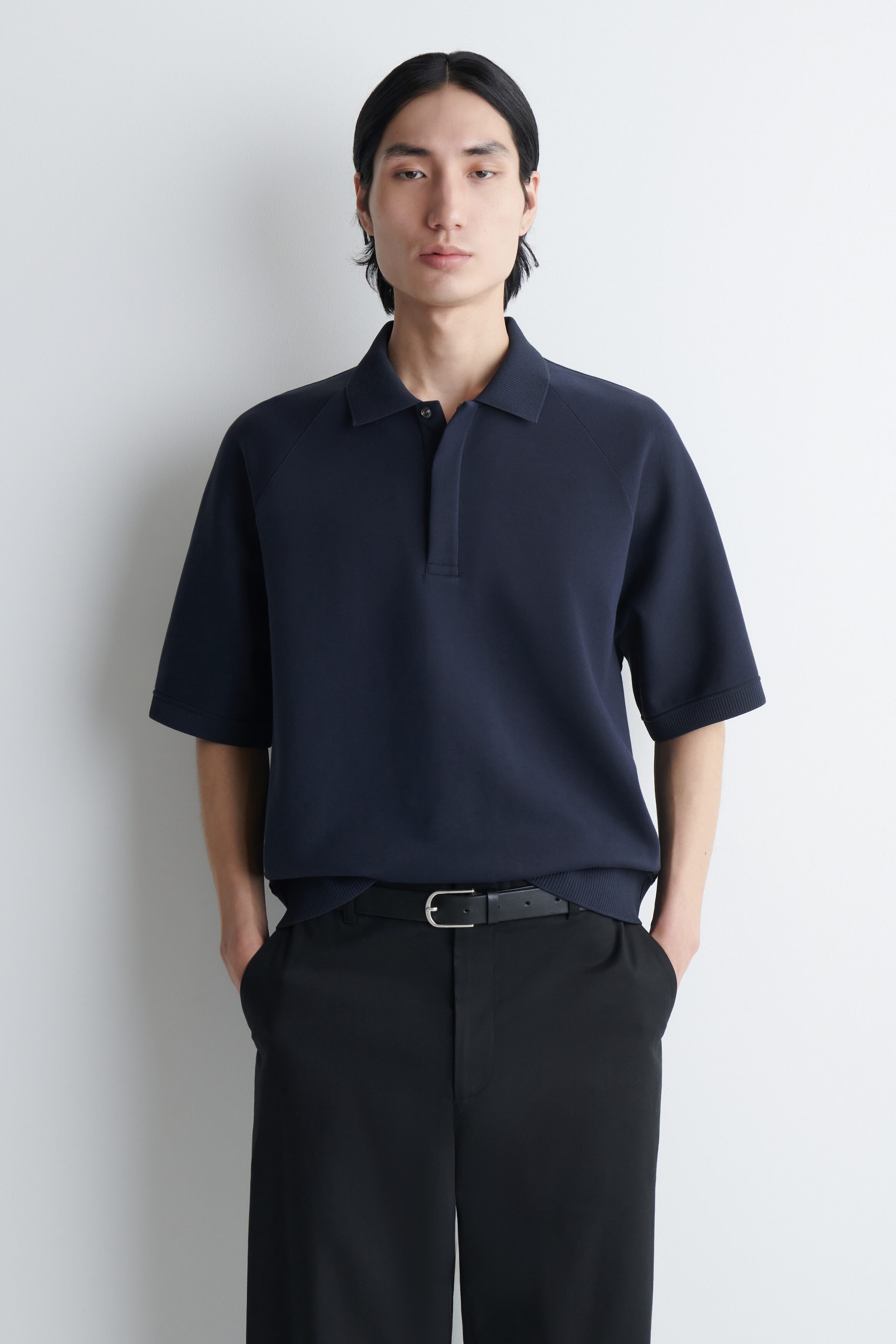 A navy blue scuba jersey polo shirt with short raglan sleeves, a classic collar, and a concealed placket is being worn by the model. The polo shirt is tucked into black trousers styled with a black belt. A navy blue scuba jersey polo shirt with short raglan sleeves, a classic collar, and a concealed placket is being worn by the model. The polo shirt is tucked into black trousers styled with a black belt.
