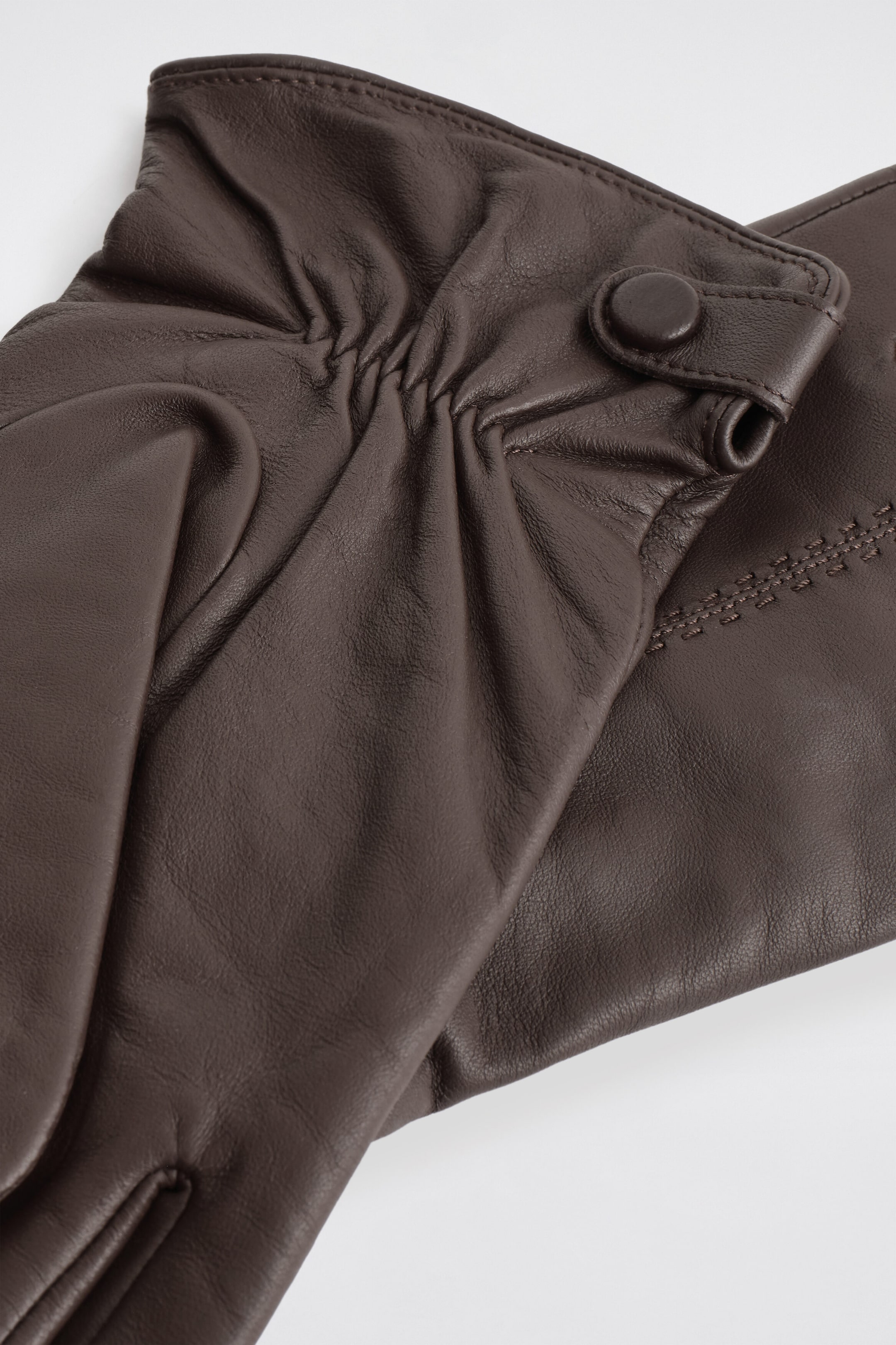 These dark brown leather gloves are resting one on top of the other, showcasing a ruched wrist with a leather-covered snap closure on the top glove and decorative tonal topstitching on the glove underneath. These dark brown leather gloves are resting one on top of the other, showcasing a ruched wrist with a leather-covered snap closure on the top glove and decorative tonal topstitching on the glove underneath.