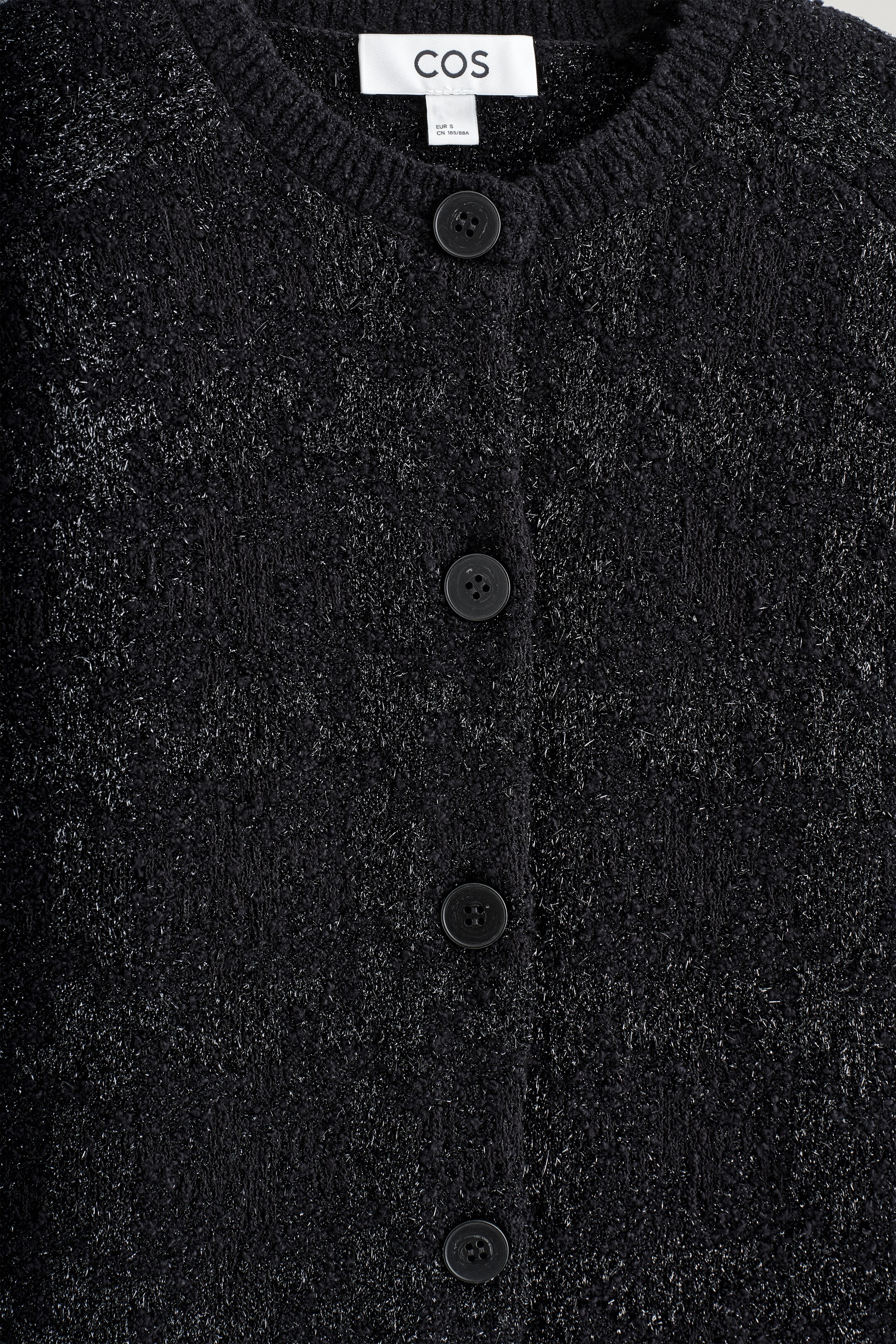 Agrandir l'image: Un cardigan en maille bouclée noire est exposé de face, avec des fils métalliques qui créent un éclat subtil sur sa surface texturée et capturent la lumière. Il est conçu avec un col rond, une fermeture à boutons sur le devant montrant quatre boutons ronds et sombres, et une étiquette blanche COS est visible à l'arrière du col.