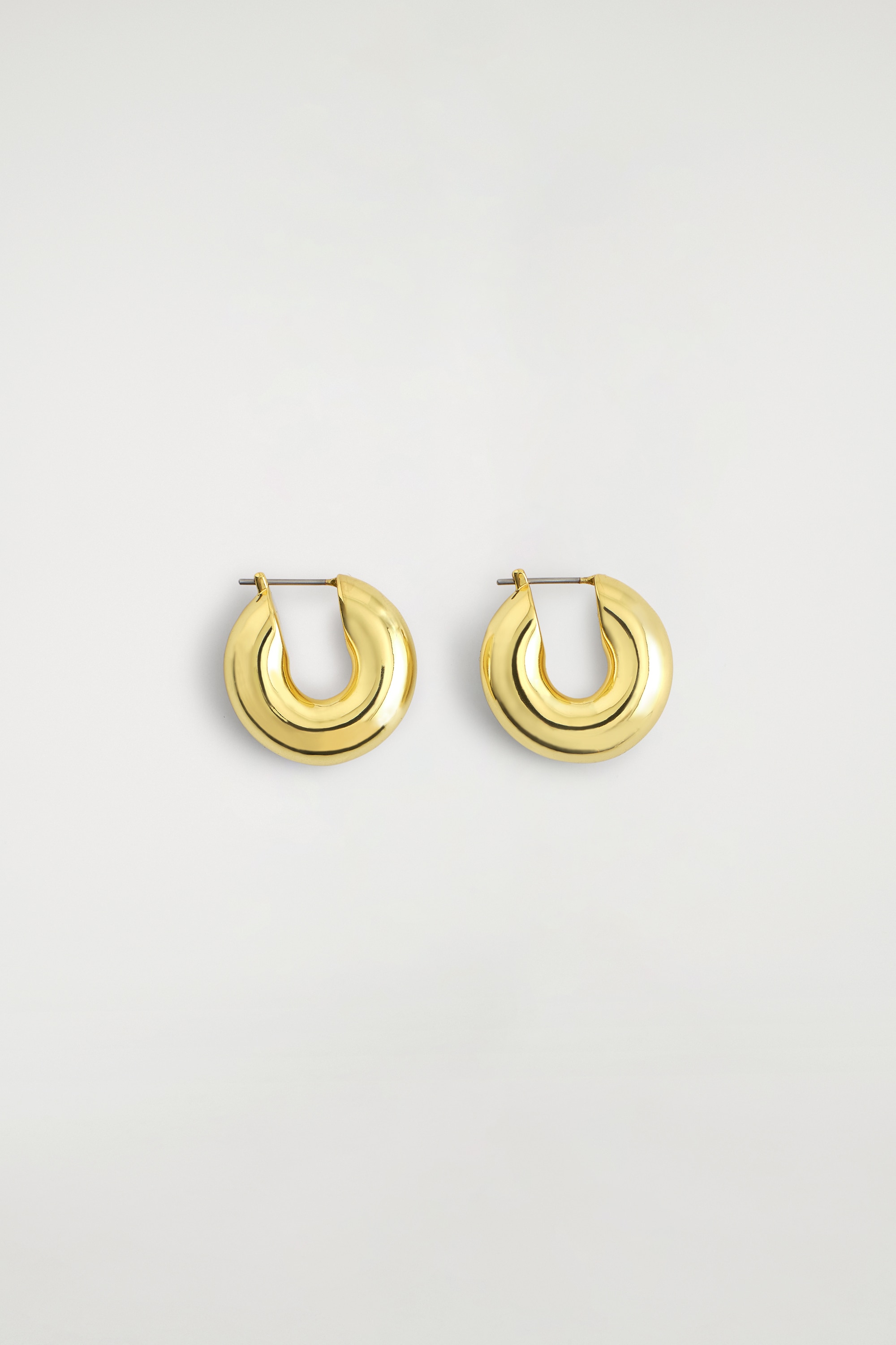 Mini brincos de argola volumosa em metal dourado polido estÃ£o a ser usados nos lÃ³bulos das orelhas, vistos de frente para mostrar o seu design espesso e arredondado e o acabamento de alto brilho.