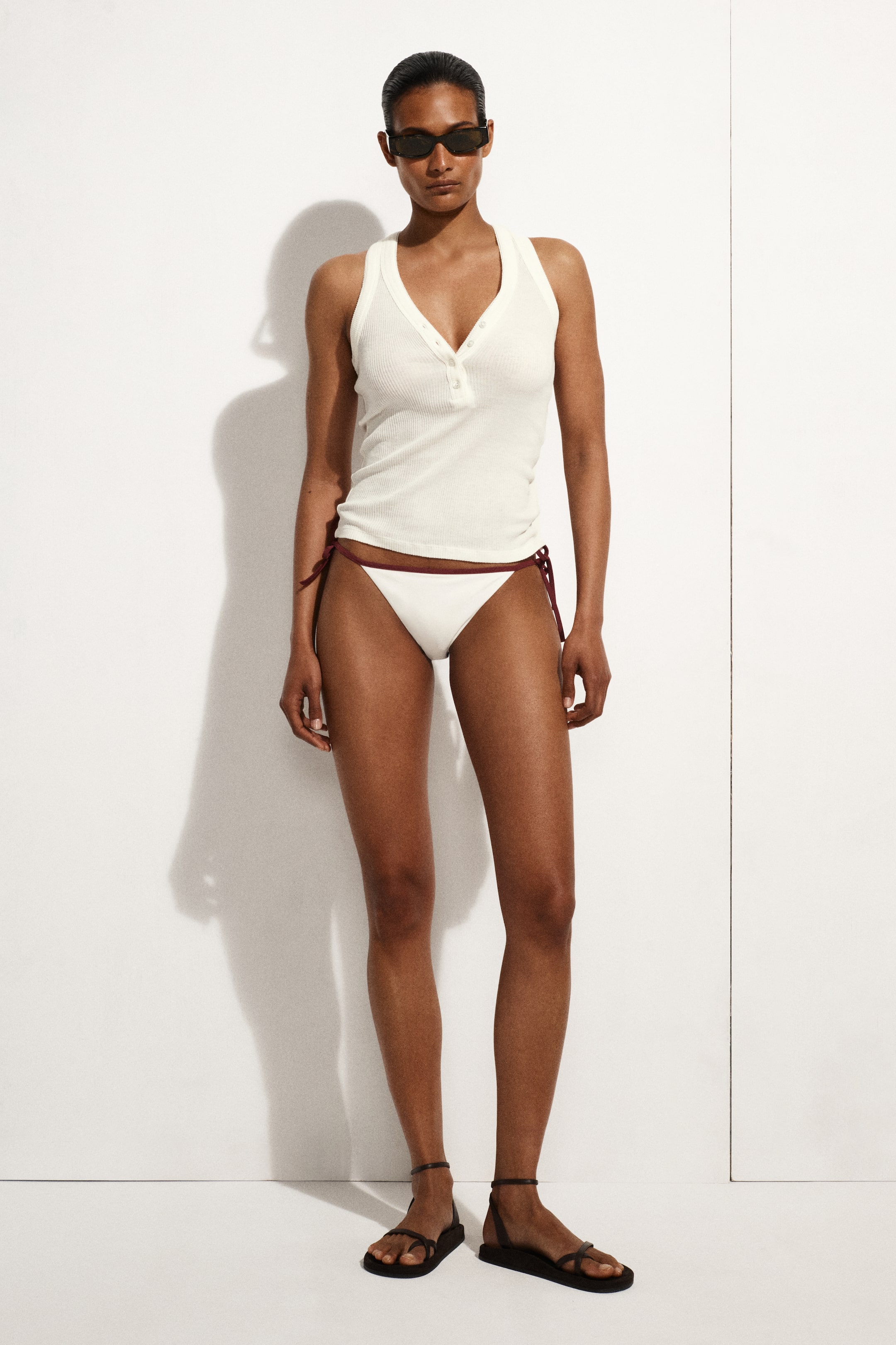 Ver imagen más grande: Una camiseta de tirantes blanca acanalada, con escote en V y una tapeta de cuatro botones, se presenta desde el frente con braguitas de bikini de cordones blancos que tienen lazos rojo oscuro. Sandalias planas negras con múltiples correas completan el conjunto.