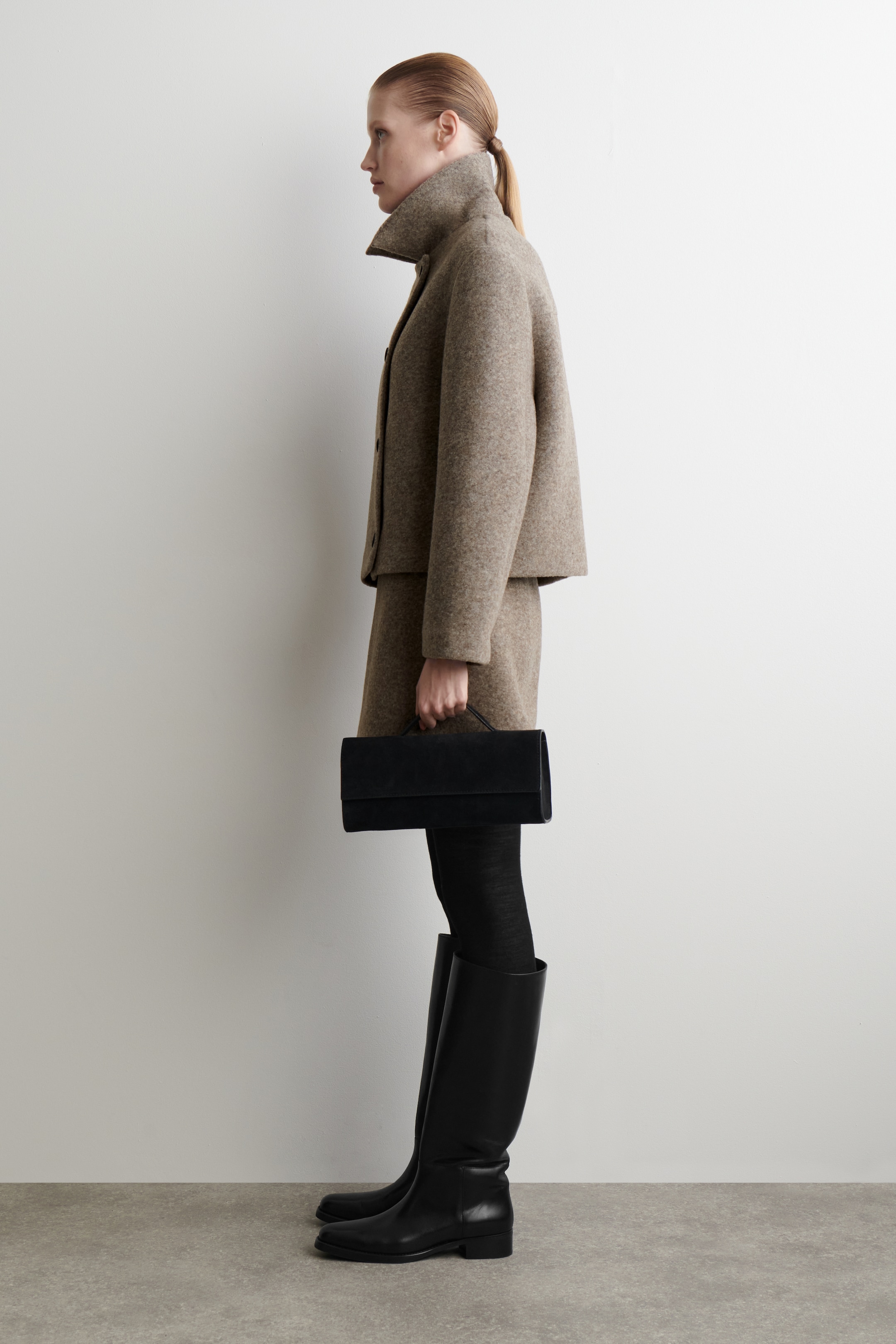Ver imagen más grande: Una chaqueta de corte ancho, color marrón claro, texturizada, con un cuello alto levantado y botones visibles se lleva con una falda en línea A a juego, vista de perfil lateral. Se sostiene un bolso rectangular negro con asa superior, y se llevan botas altas de cuero negro sobre medias opacas negras.