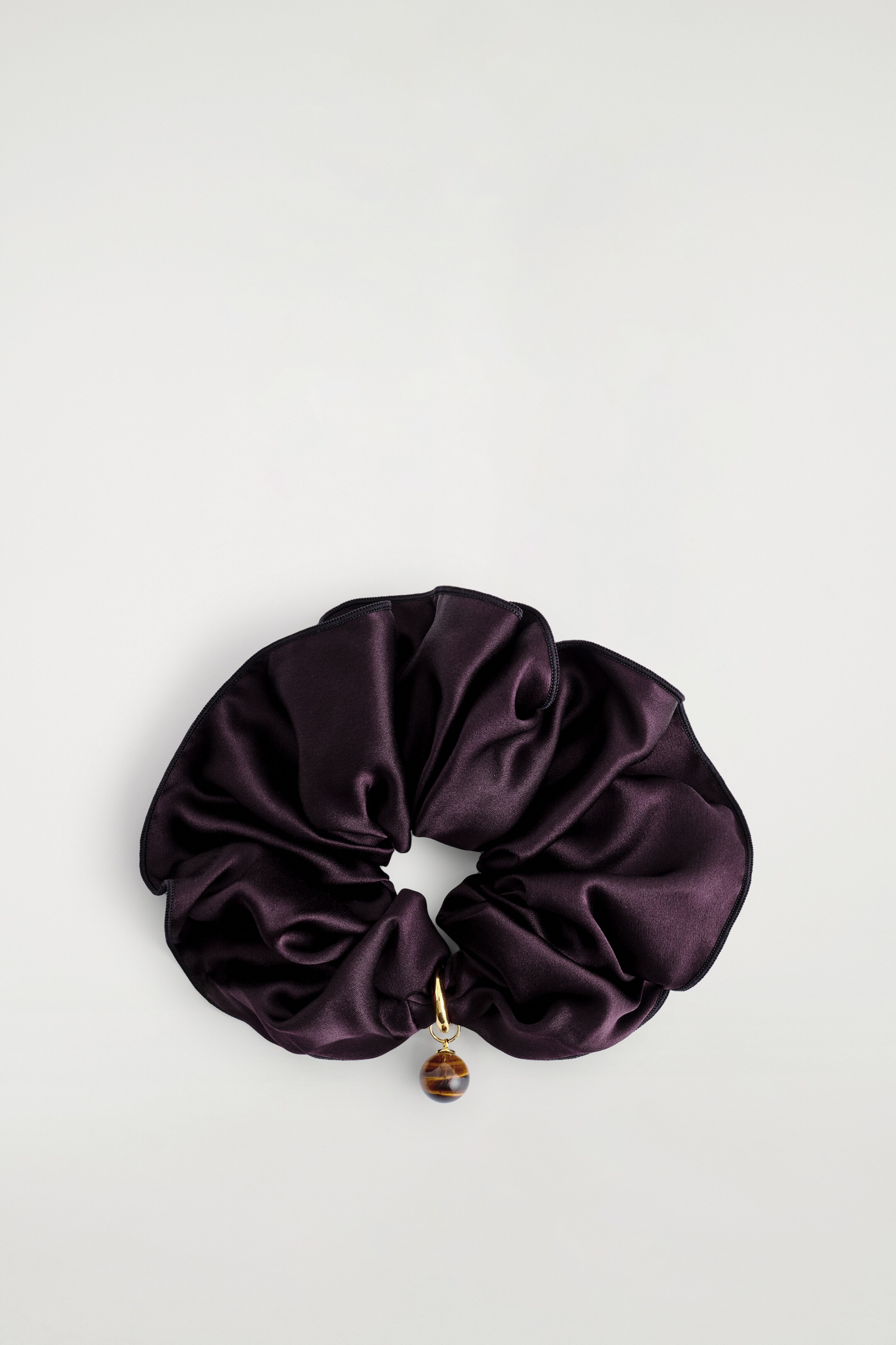 Visualizza immagine più grande: Un voluminoso scrunchie, visto come TOP, mostra una seta color prugna scuro lucente mulberry. Un charm sferico in occhio di tigre, attaccato con un anello tono oro, sta pendendo dallo scrunchie.