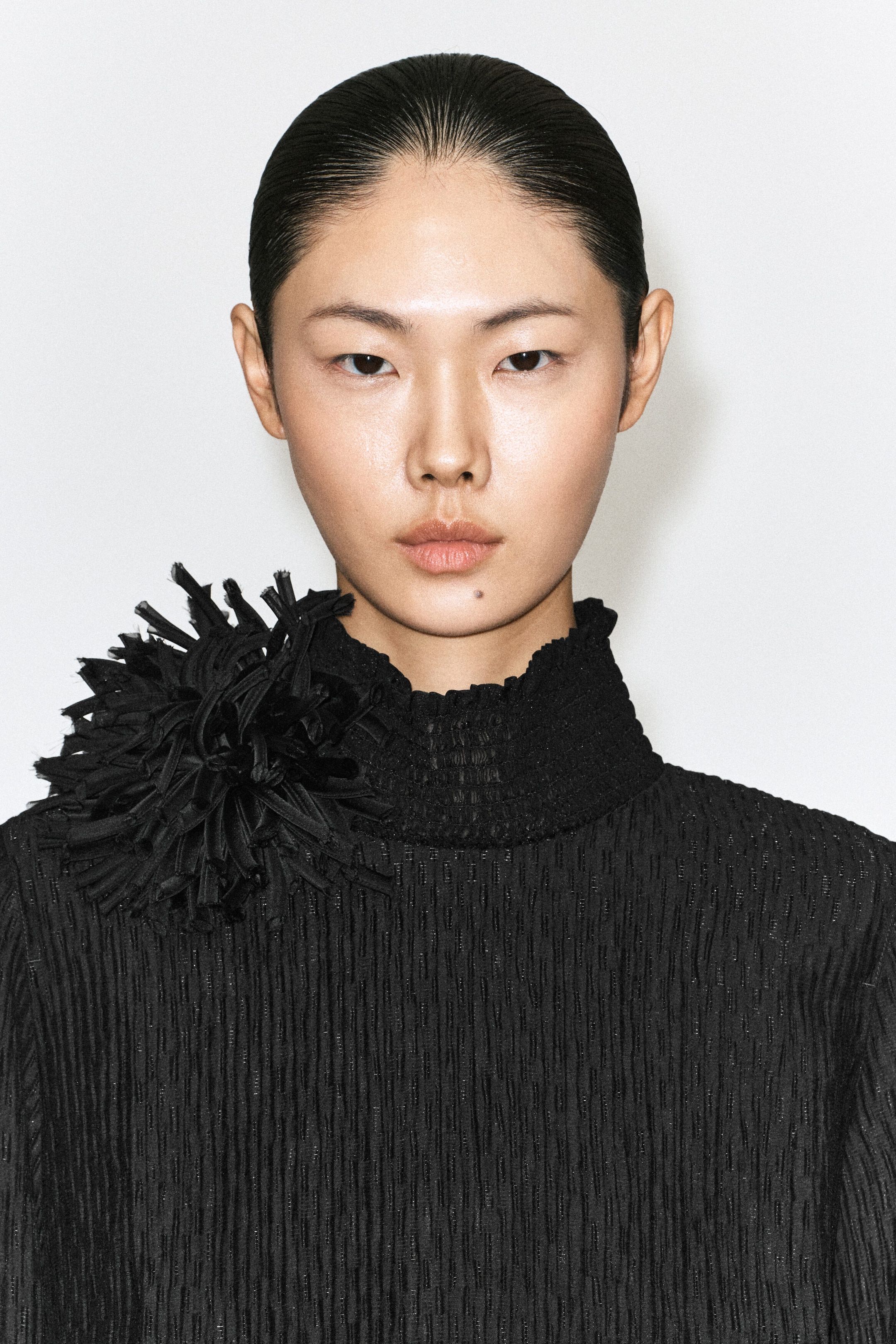 Une broche rosette florale noire, ornée de pétales en tissu denses et pointus, est portée à l’épaule par le mannequin sur un haut noir texturé à col montant faux. Une broche rosette florale noire, ornée de pétales en tissu denses et pointus, est portée à l’épaule par le mannequin sur un haut noir texturé à col montant faux.
