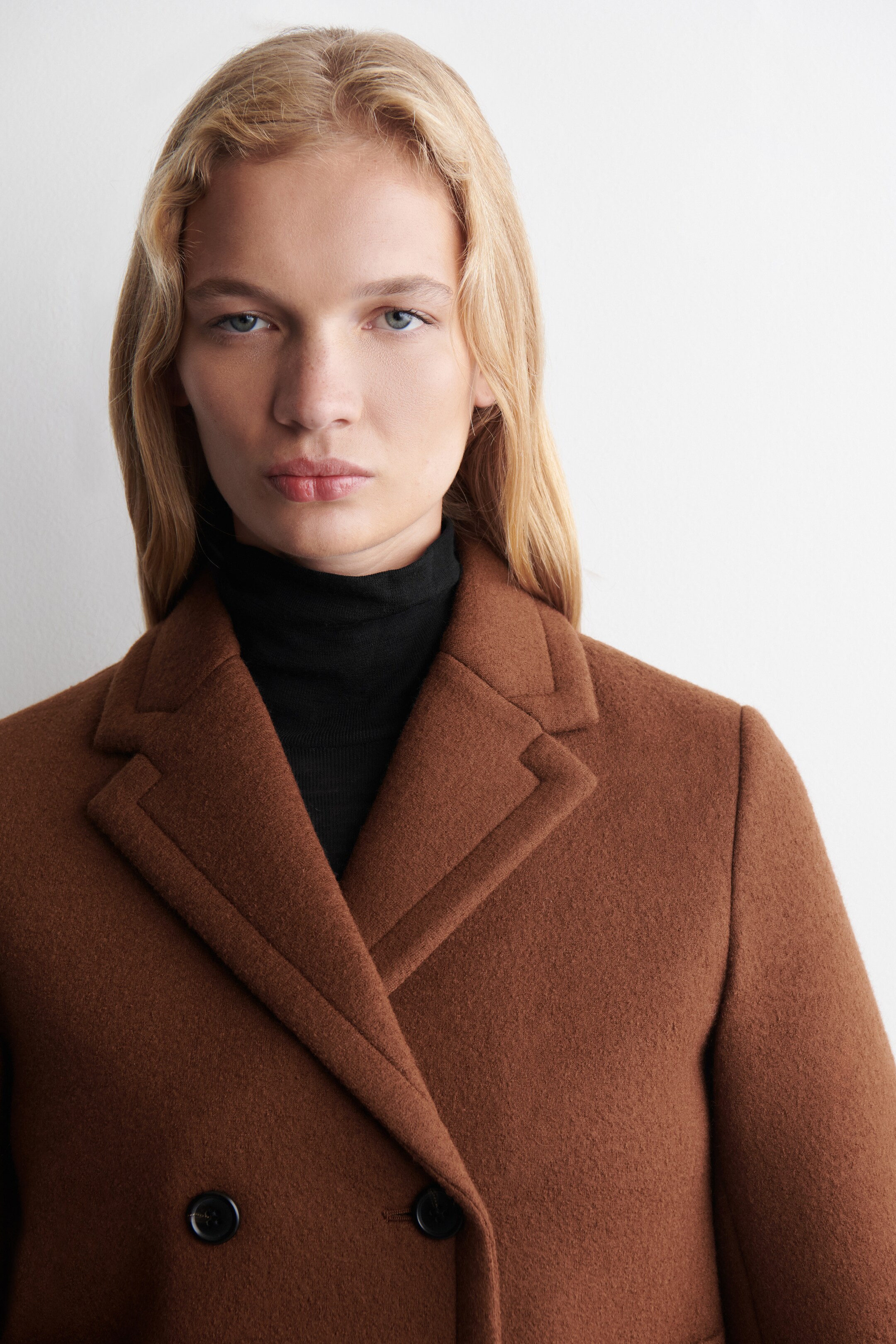View larger image: A brown double-breasted coat is being shown in a front-facing view, featuring prominent notch lapels and two black buttons on its textured fabric. A black turtleneck is layering underneath the coat, filling the neckline.