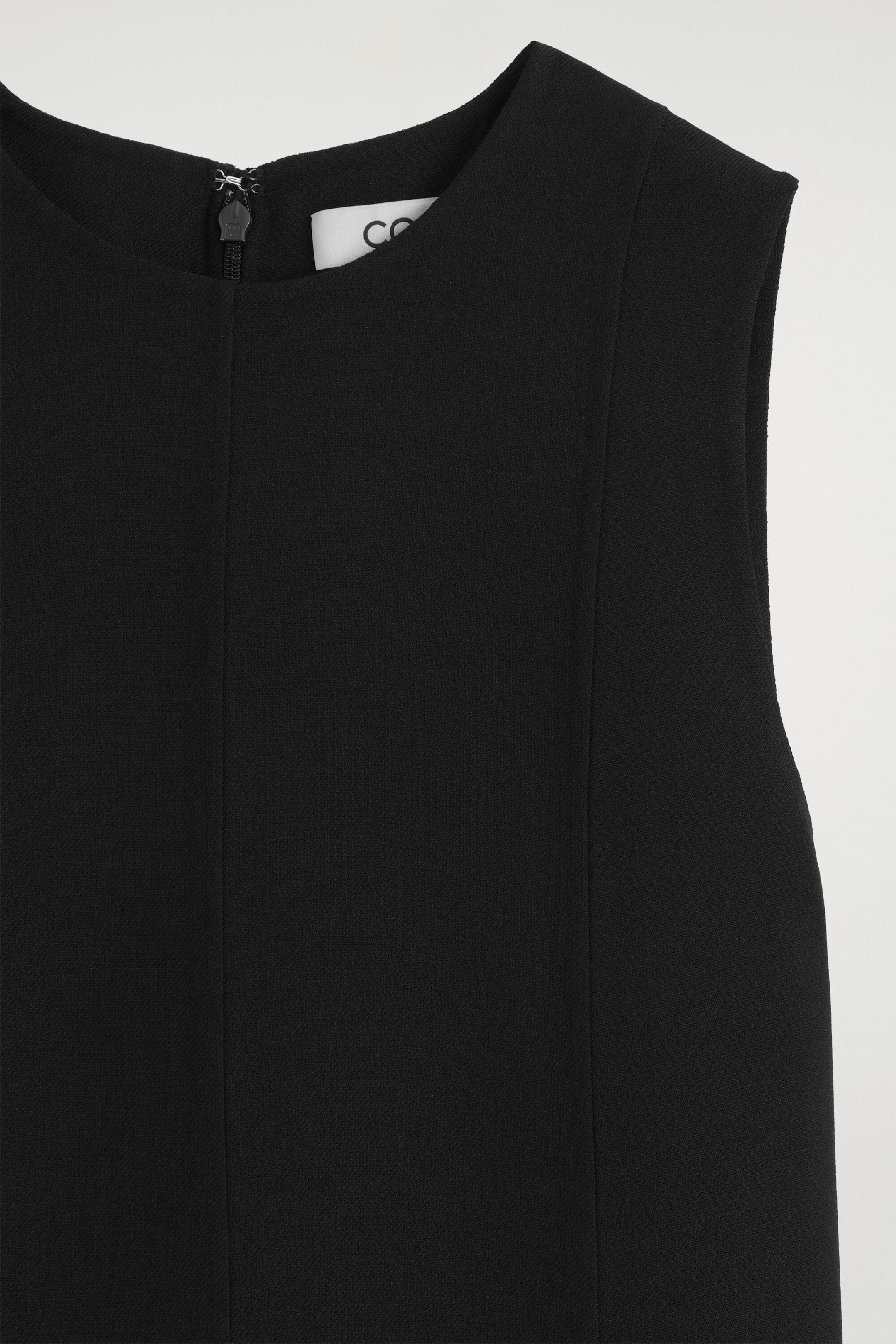 A black sleeveless garment is shown in close-up, featuring a round neckline, structured vertical panel seams, and a zip fastening with a black pull tab at the centre of the collar. A black sleeveless garment is shown in close-up, featuring a round neckline, structured vertical panel seams, and a zip fastening with a black pull tab at the centre of the collar.