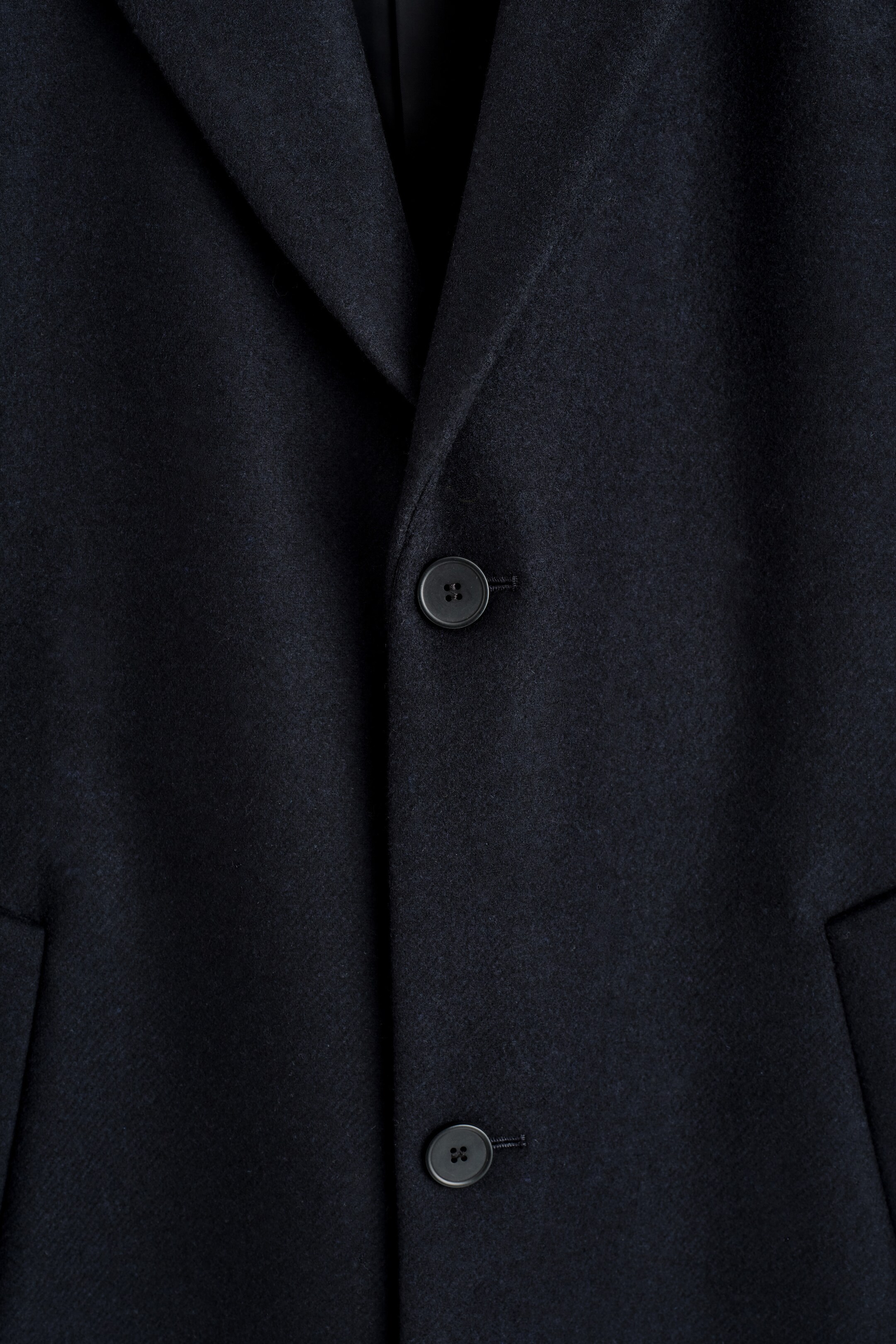 View larger image: A dark, single-breasted coat is presenting its FRONT, displaying classic notch lapels and two dark, round buttons on the closure. A welt pocket is also partially visible on the lower left side.