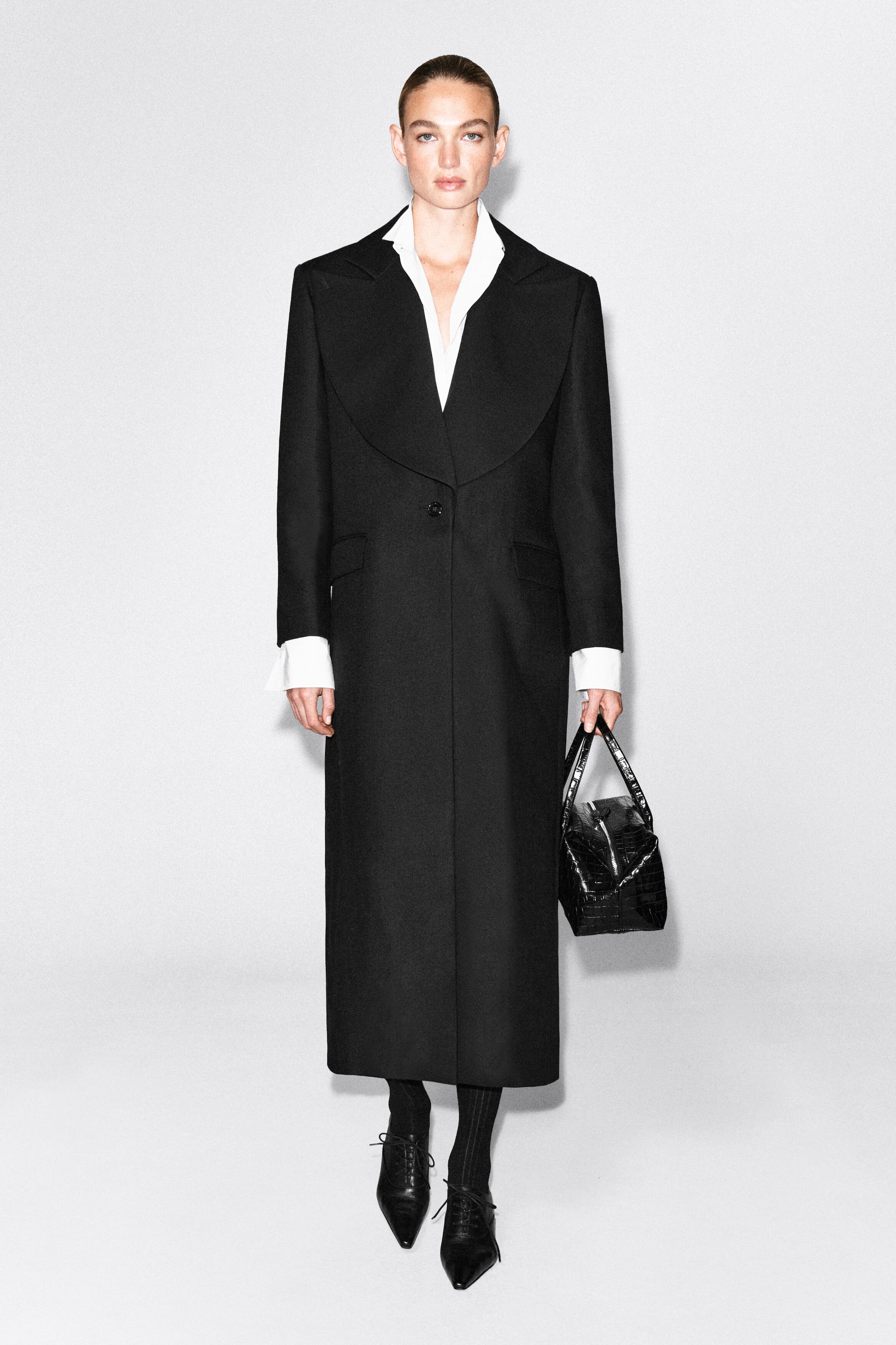 A long black single-breasted coat featuring wide, draped lapels and a single-button fastening is worn by the model. The coat is layered over a white collared shirt and styled with black lace-up shoes and a black textured handbag held by the model.