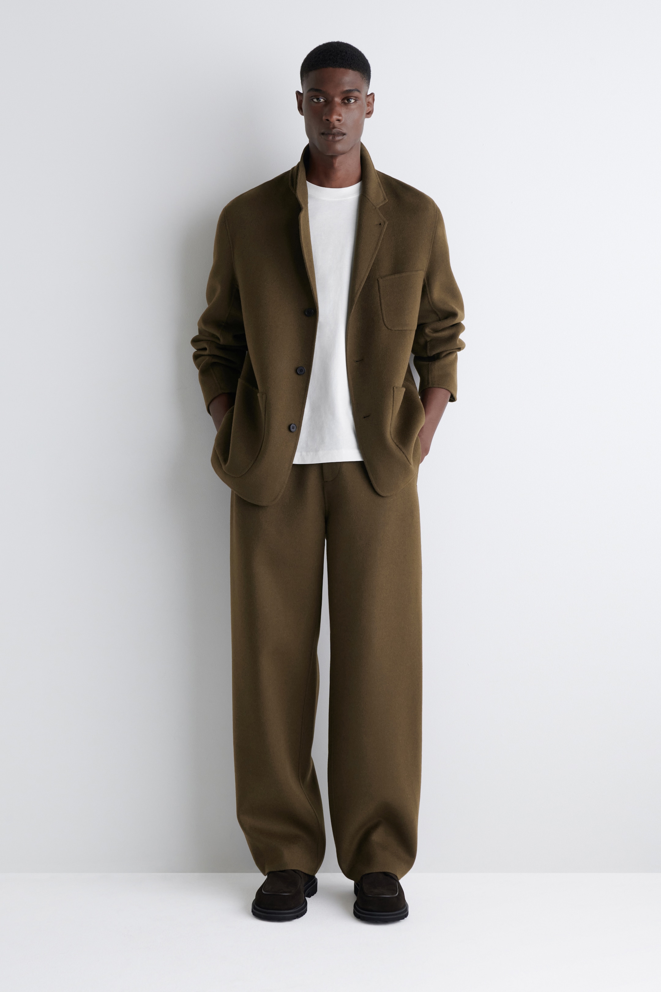 Agrandir l'image: Un costume deux pièces vert olive foncé orienté vers l'avant est présenté, composé d'une veste déstructurée à coupe décontractée avec un col montant souple, deux boutons sombres et trois poches plaquées, portée sur un t-shirt blanc à col rond. Le pantalon assorti à jambes larges s'accumule sur des chaussures à enfiler sombres à semelles épaisses.