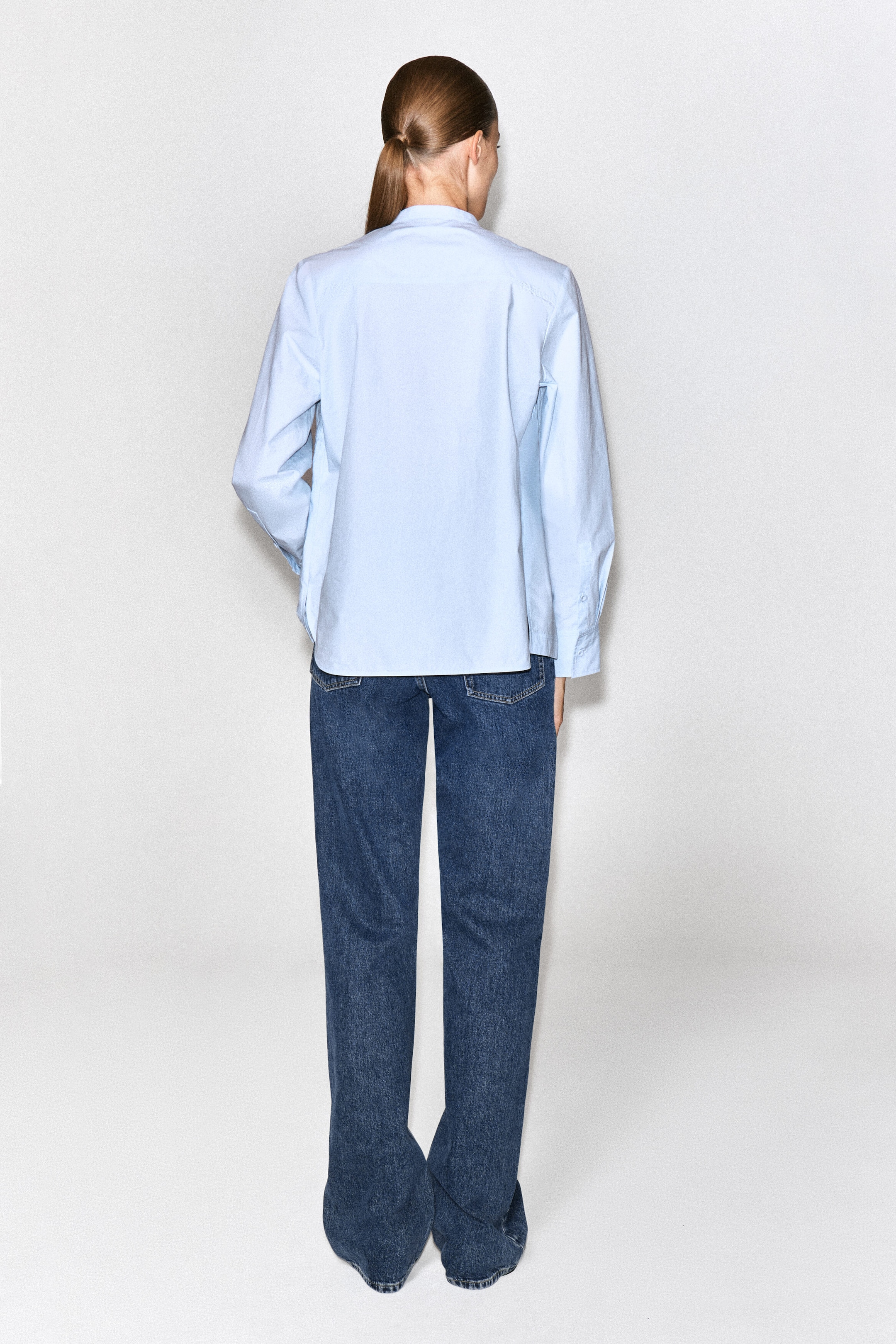 The model is wearing a light blue cotton blouse styled with dark blue straight-leg jeans. The long-sleeved blouse is shown from the back, highlighting its relaxed fit, classic collar and straight hem. The model is wearing a light blue cotton blouse styled with dark blue straight-leg jeans. The long-sleeved blouse is shown from the back, highlighting its relaxed fit, classic collar and straight hem.