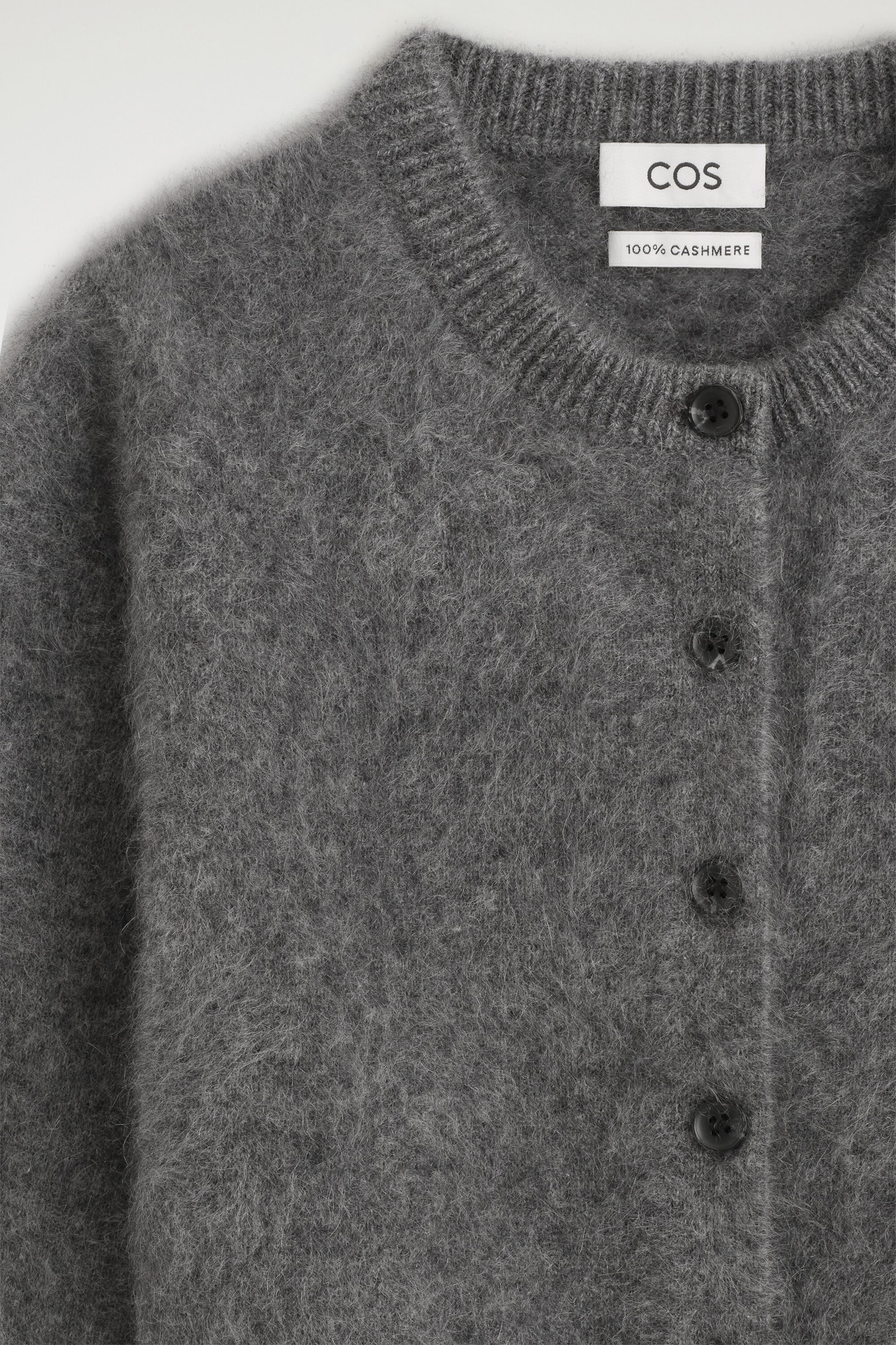 A kids' brushed pure cashmere cardigan is shown in a close-up front view, highlighting its soft, fuzzy grey mélange texture. The cardigan features a ribbed crew neckline and dark round buttons along the front placket.