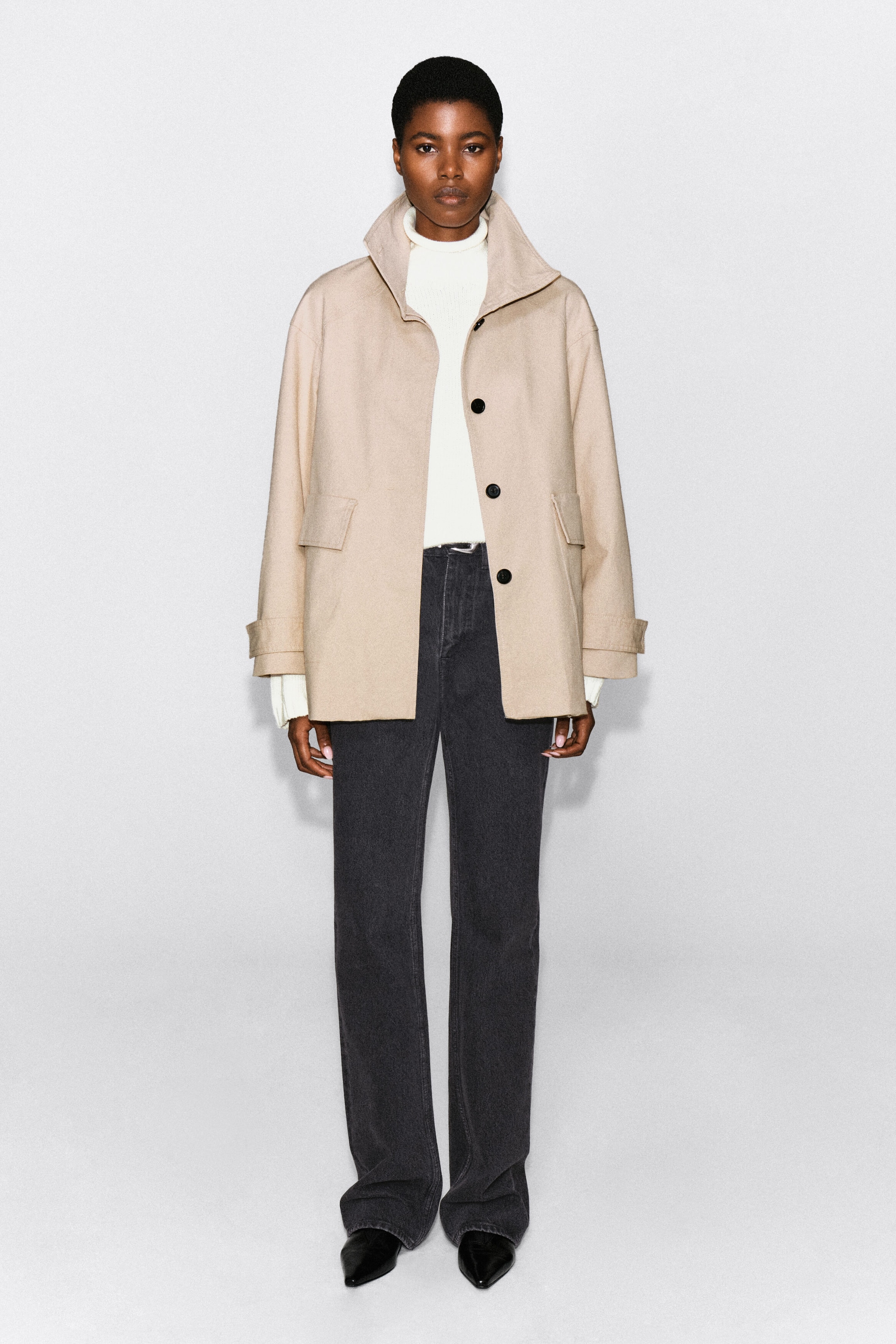A beige cotton-twill funnel-neck jacket with large dark buttons and side flap pockets is being worn by the model. The jacket is layered over a white high-neck sweater and paired with dark grey trousers. A beige cotton-twill funnel-neck jacket with large dark buttons and side flap pockets is being worn by the model. The jacket is layered over a white high-neck sweater and paired with dark grey trousers.