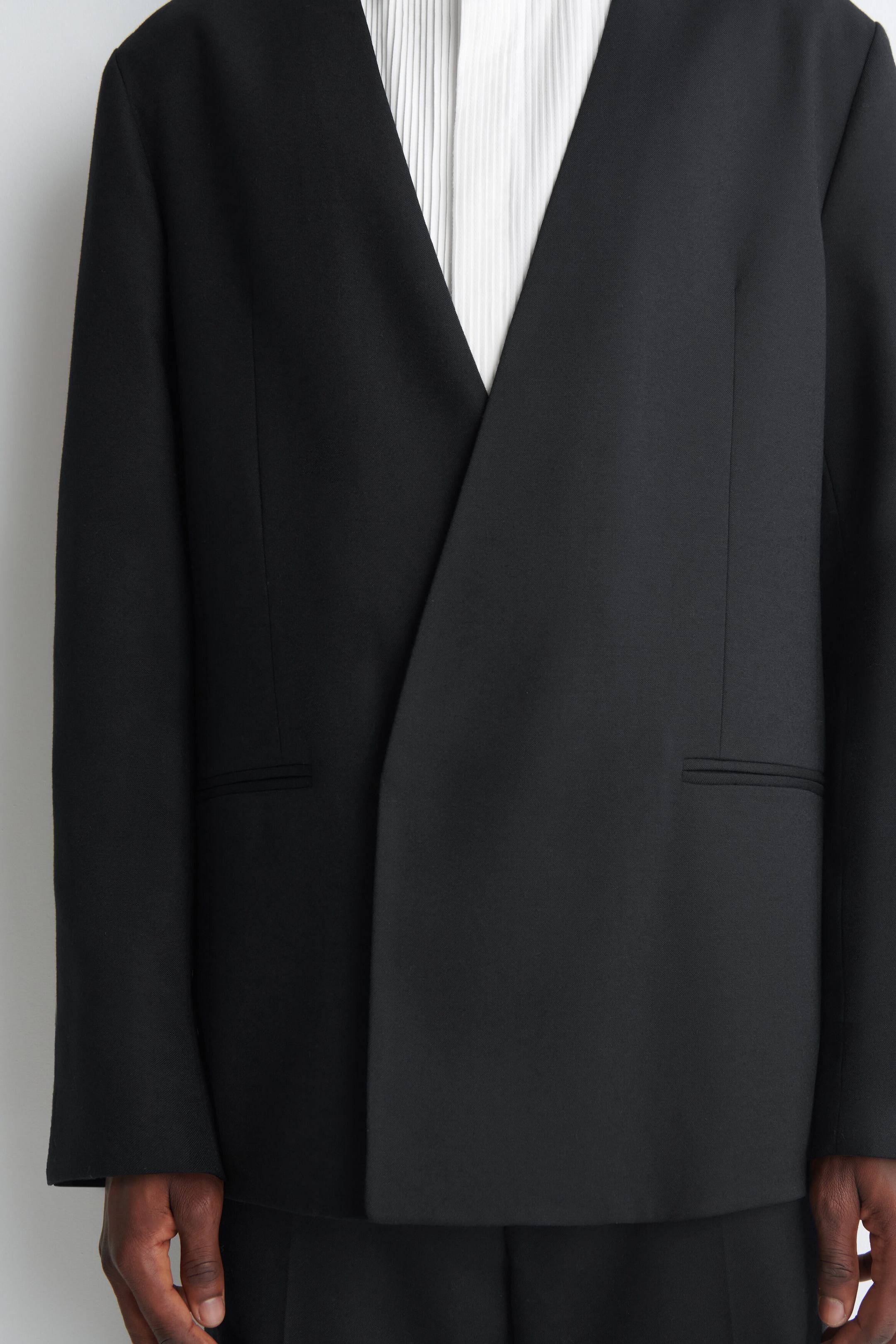 The model is wearing a black collarless blazer with an asymmetrical crossover front and welt pockets. The blazer is layered over a white pleated shirt and styled with matching black trousers. The model is wearing a black collarless blazer with an asymmetrical crossover front and welt pockets. The blazer is layered over a white pleated shirt and styled with matching black trousers.
