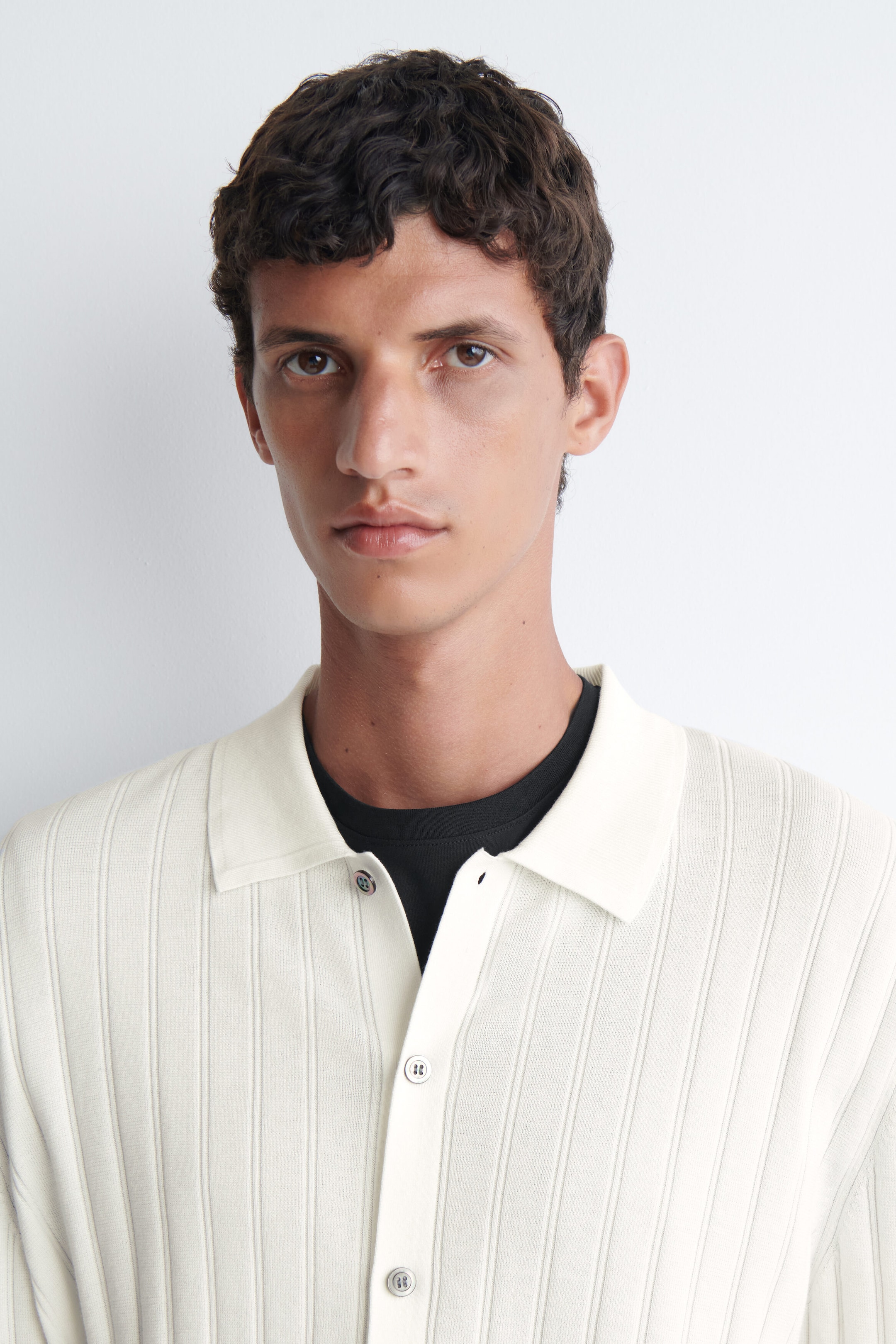 Ein gerippt gestricktes Baumwollhemd in Off-White wird vom Model getragen und verfügt über einen Kragen sowie eine durchgehende Knopfleiste vorn. Das Hemd ist über einem schwarzen T-Shirt mit Rundhalsausschnitt getragen. Ein gerippt gestricktes Baumwollhemd in Off-White wird vom Model getragen und verfügt über einen Kragen sowie eine durchgehende Knopfleiste vorn. Das Hemd ist über einem schwarzen T-Shirt mit Rundhalsausschnitt getragen.