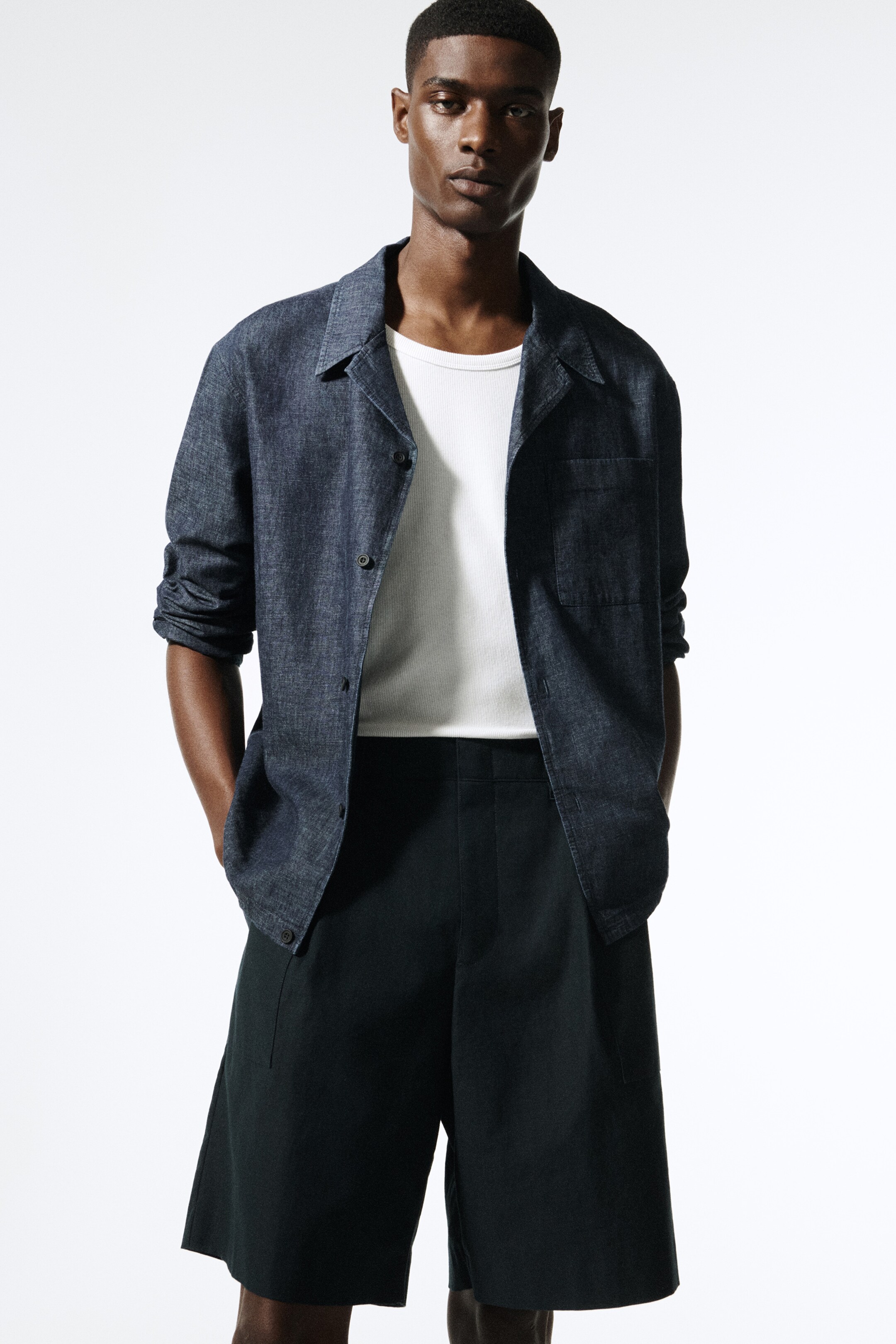 Größeres Bild anzeigen: Ein dunkelblaues Overshirt mit Resortkragen, Knopfverschluss und einer Brusttasche wird offen über einem weißen gerippten T-Shirt getragen. Es wird mit dunkel gefärbten, plissierten Chino-Shorts kombiniert, die eine entspannte Passform vorne bieten.
