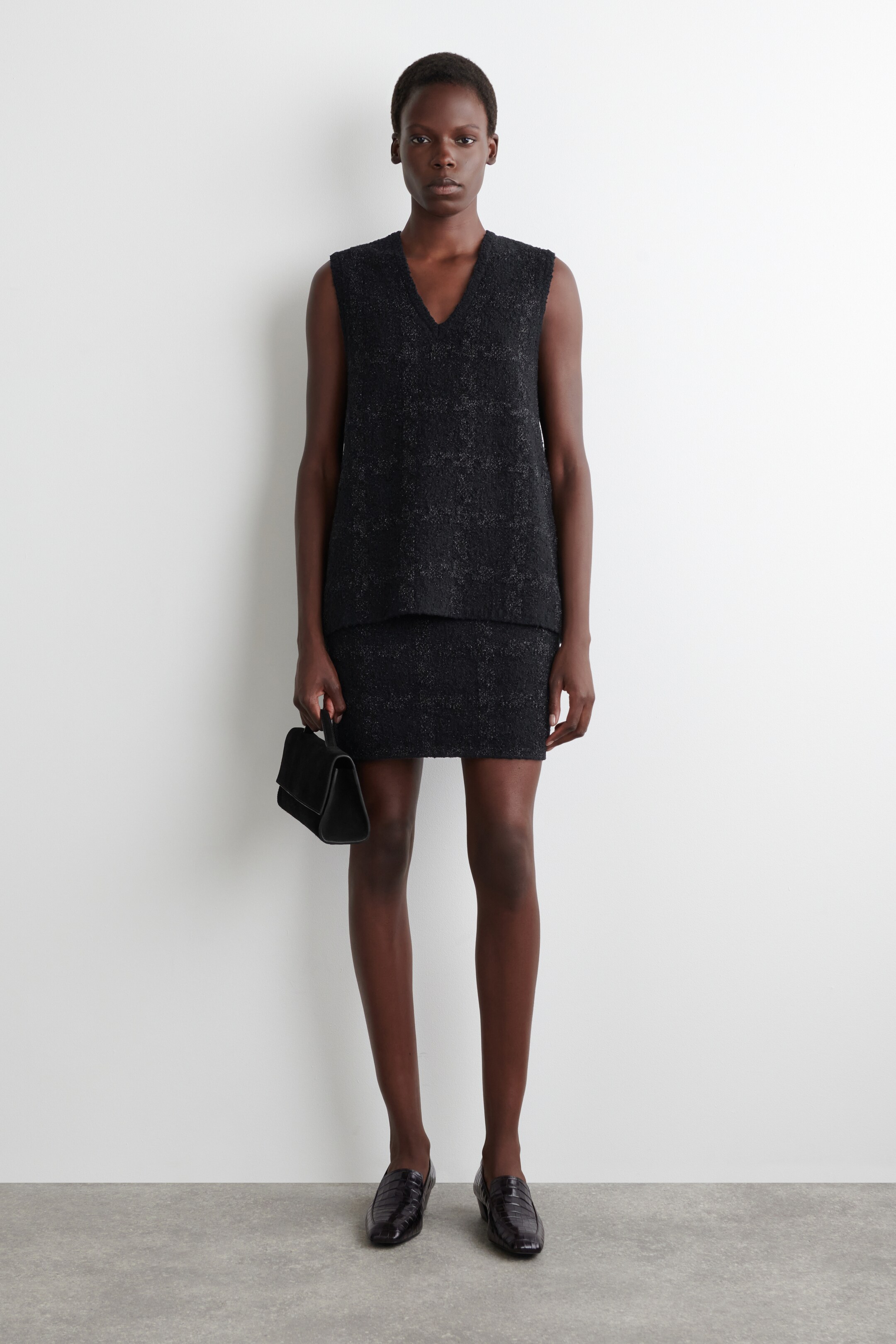 Agrandir l'image: Un haut noir texturé sans manches avec un décolleté en V et une mini-jupe assortie sont présentés de face. Le haut est légèrement ample et la jupe s'arrête au-dessus du genou, tandis qu'un petit sac à main noir structuré est tenu sur le côté.
