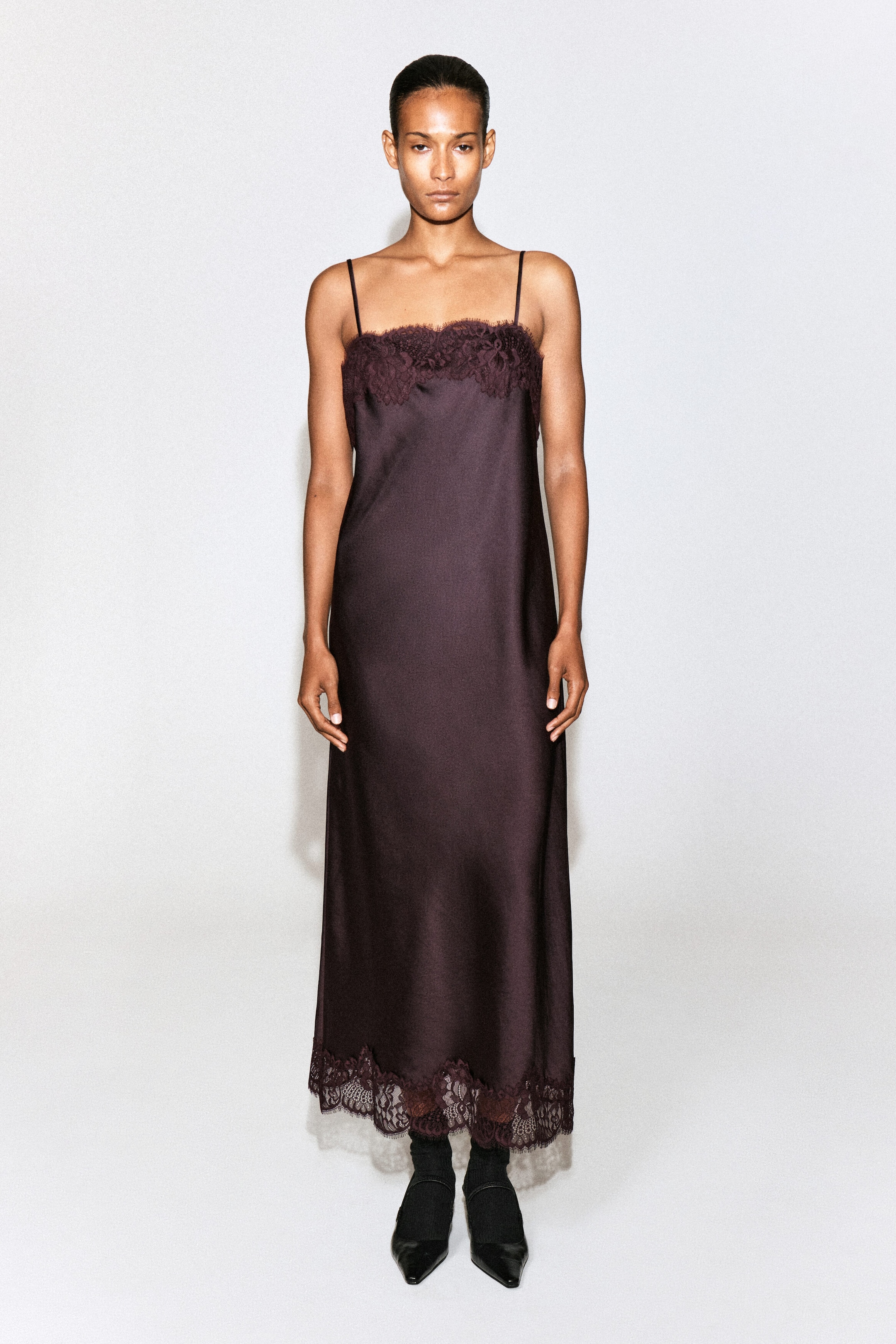 Ein dunkelpflaumenfarbenes Satin-Slipkleid wird vom Model getragen und verfügt über schmale Spaghettiträger, einen geraden Ausschnitt mit Spitzenbesatz und einen passenden Spitzenabschluss am Saum. Das midi-lange Kleid ist mit schwarzen Socken und spitz zulaufenden Schuhen kombiniert.