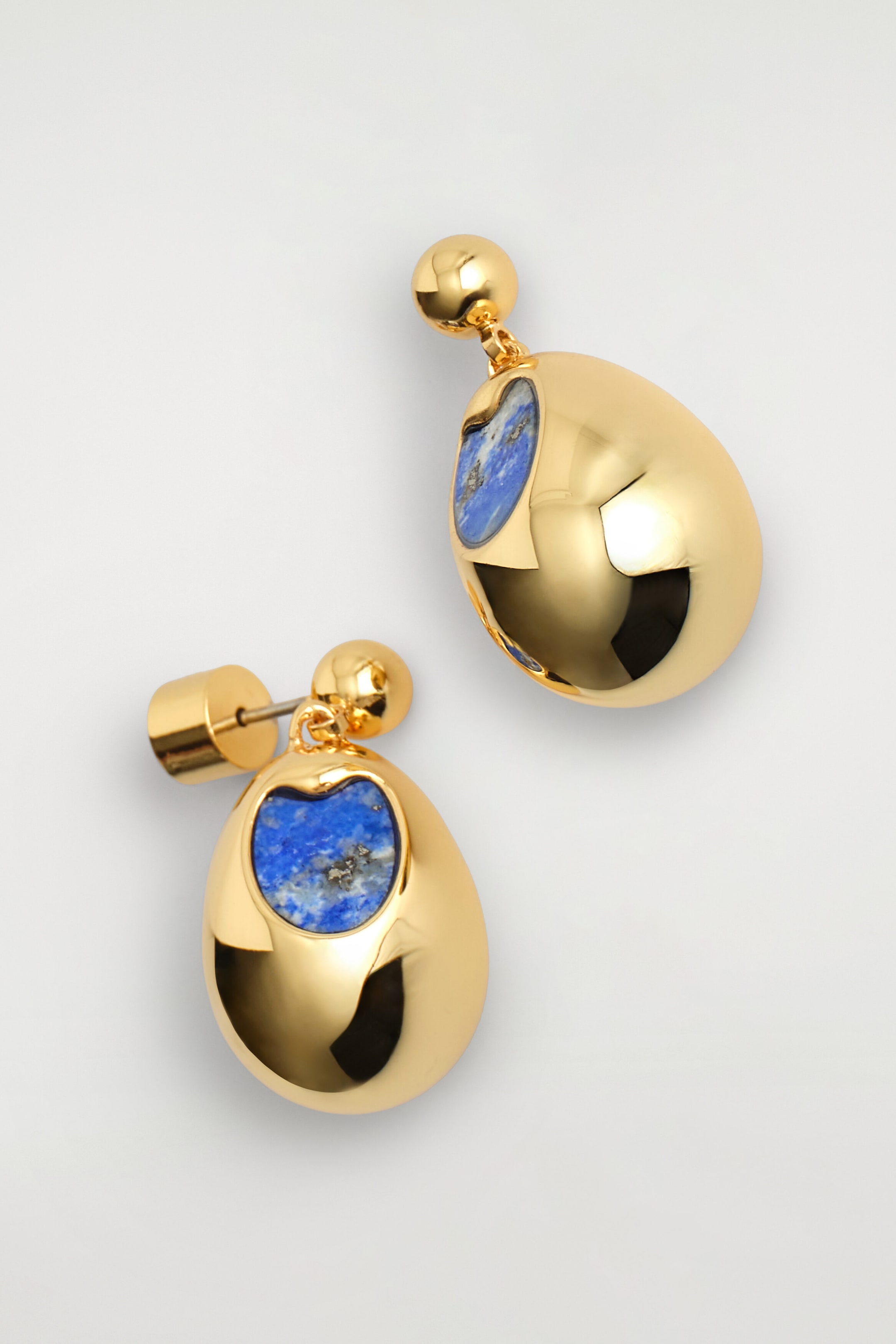 Les boucles d’oreilles pendantes à détail de pierre affichent une finition polie ton or, avec un pendentif en forme de goutte bulbeuse suspendu à un petit clou sphérique. Chaque boucle d’oreille présente, à l’avant, un incrustation de pierre bleue aux contours organiques et au veinage naturel, l’une d’elles étant positionnée de manière à montrer sa tige et son fermoir cylindrique.