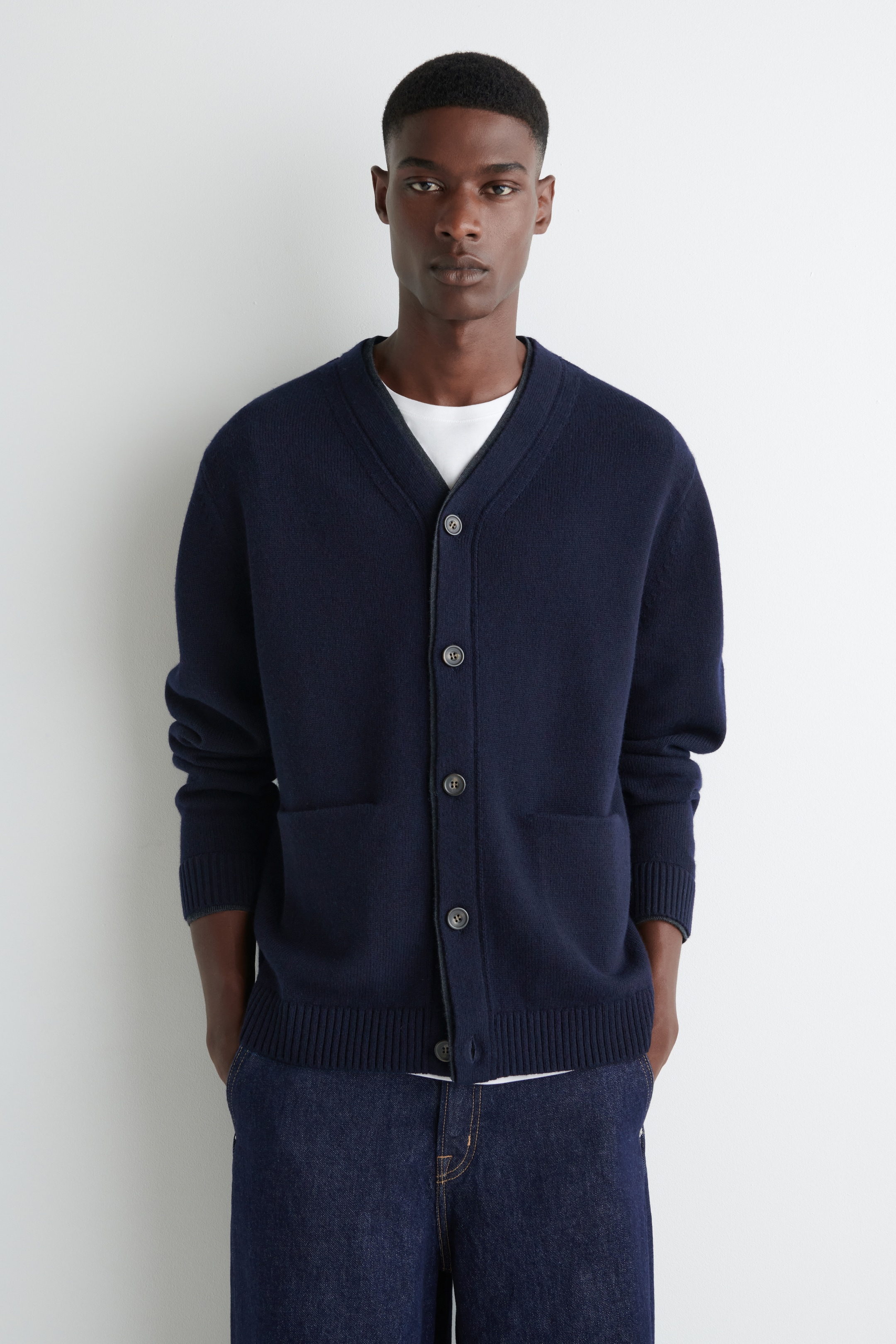 Grösseres Bild ansehen: Ein marineblauer Cardigan wird präsentiert, der über einen V-Ausschnitt und fünf dunkle Knöpfe verfügt, die entlang einer kontrastierenden, dunkel eingefassten Knopfleiste verlaufen. Er zeigt zwei aufgesetzte Fronttaschen, gerippte Bündchen und einen gerippten Saum und wird über einem weißen T-Shirt gestylt.
