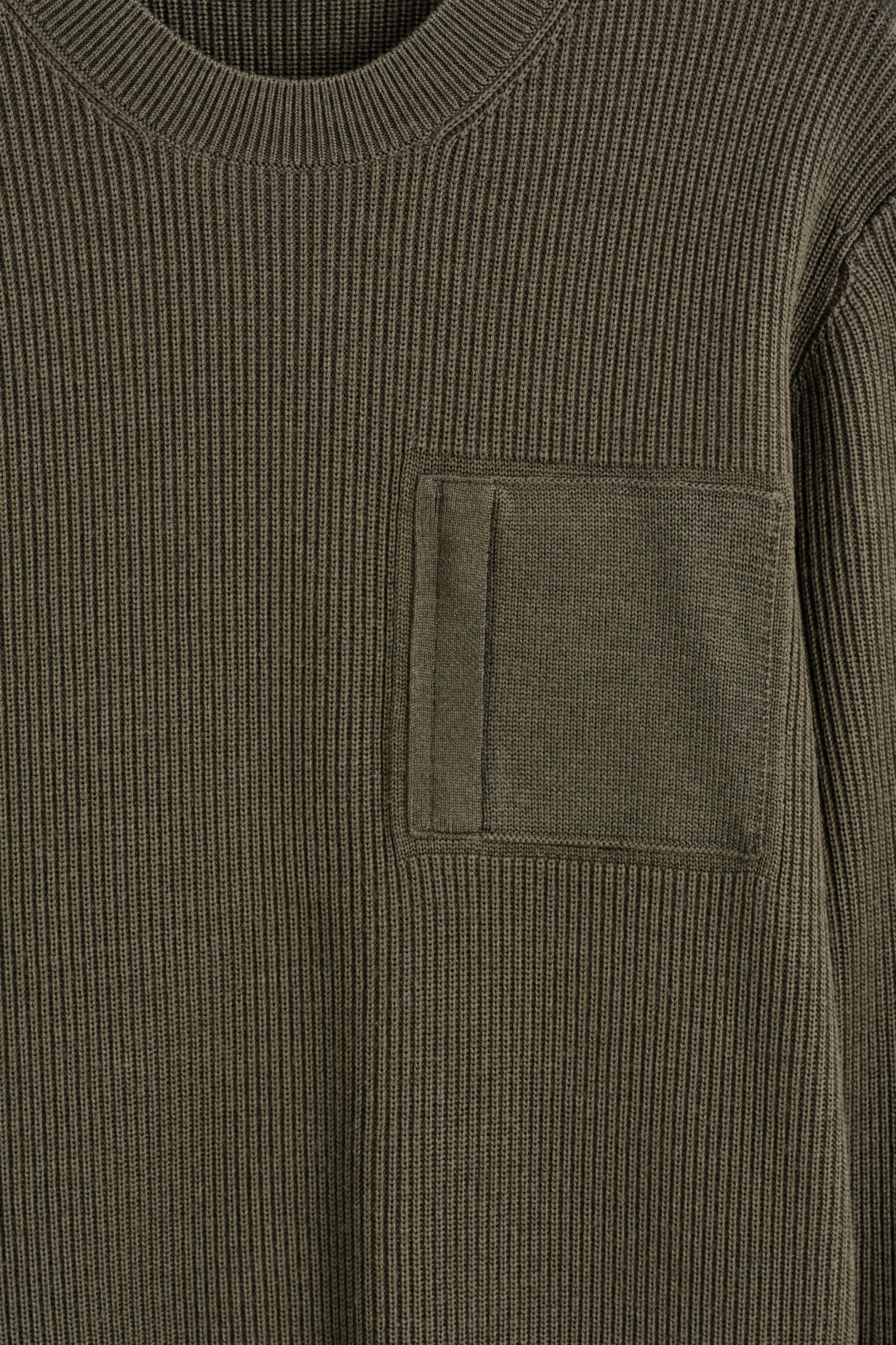 Agrandir l'image: Un pull en tricot vert olive affiche son devant, avec un motif de tricot à côtes verticales proéminent et un col rond côtelé. Une poche plaquée rectangulaire est visible sur la poitrine gauche, détaillée avec une bande de tissu verticale sur son côté gauche.