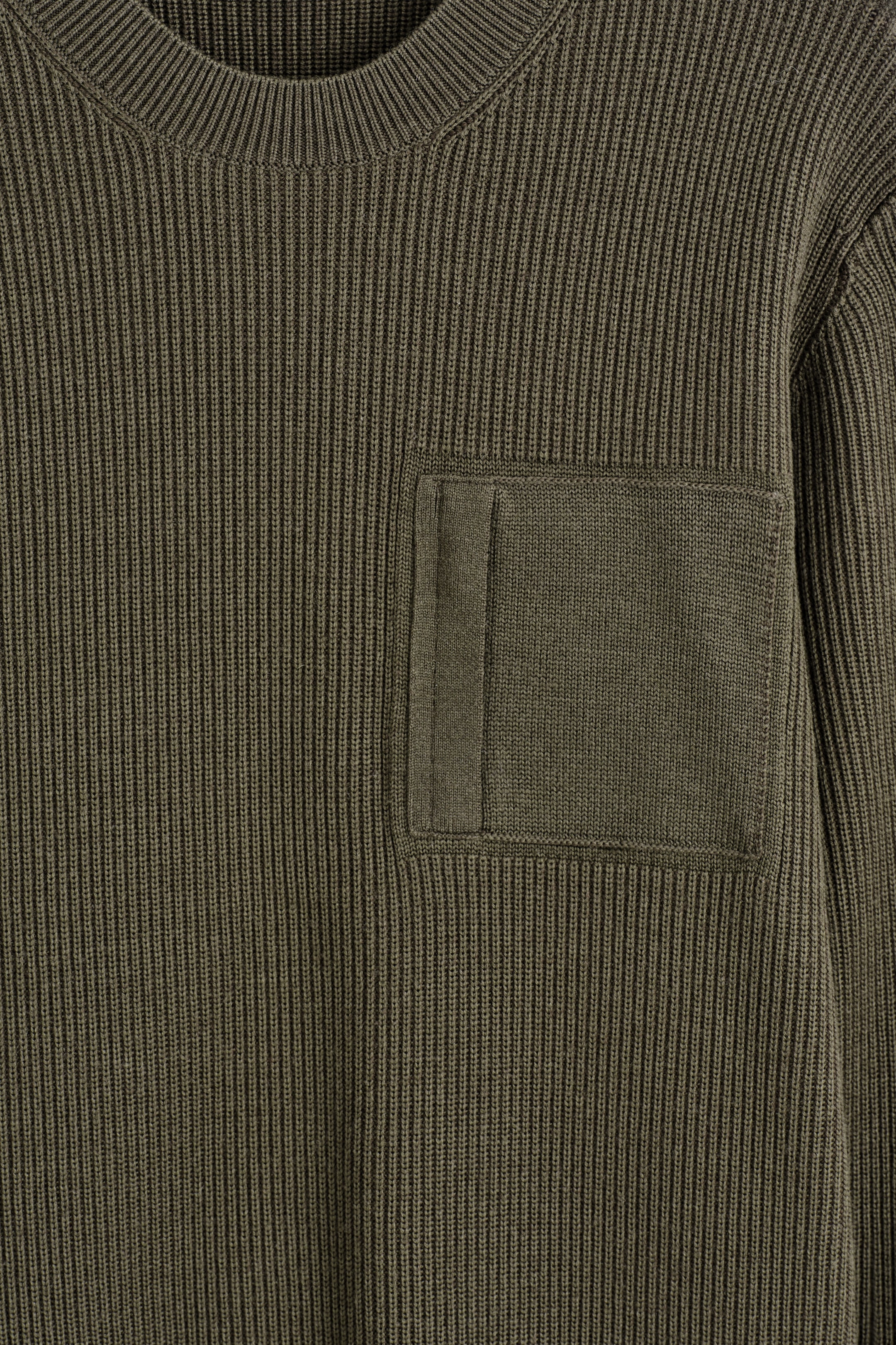 Grösseres Bild ansehen: Ein olivgrüner Strickpullover zeigt seine Vorderseite, mit einem markanten vertikalen Rippenstrickmuster und einem gerippten Rundhalsausschnitt. Eine rechteckige aufgesetzte Tasche ist auf der linken Brust sichtbar, detailliert mit einem vertikalen Stoffstreifen auf ihrer linken Seite.