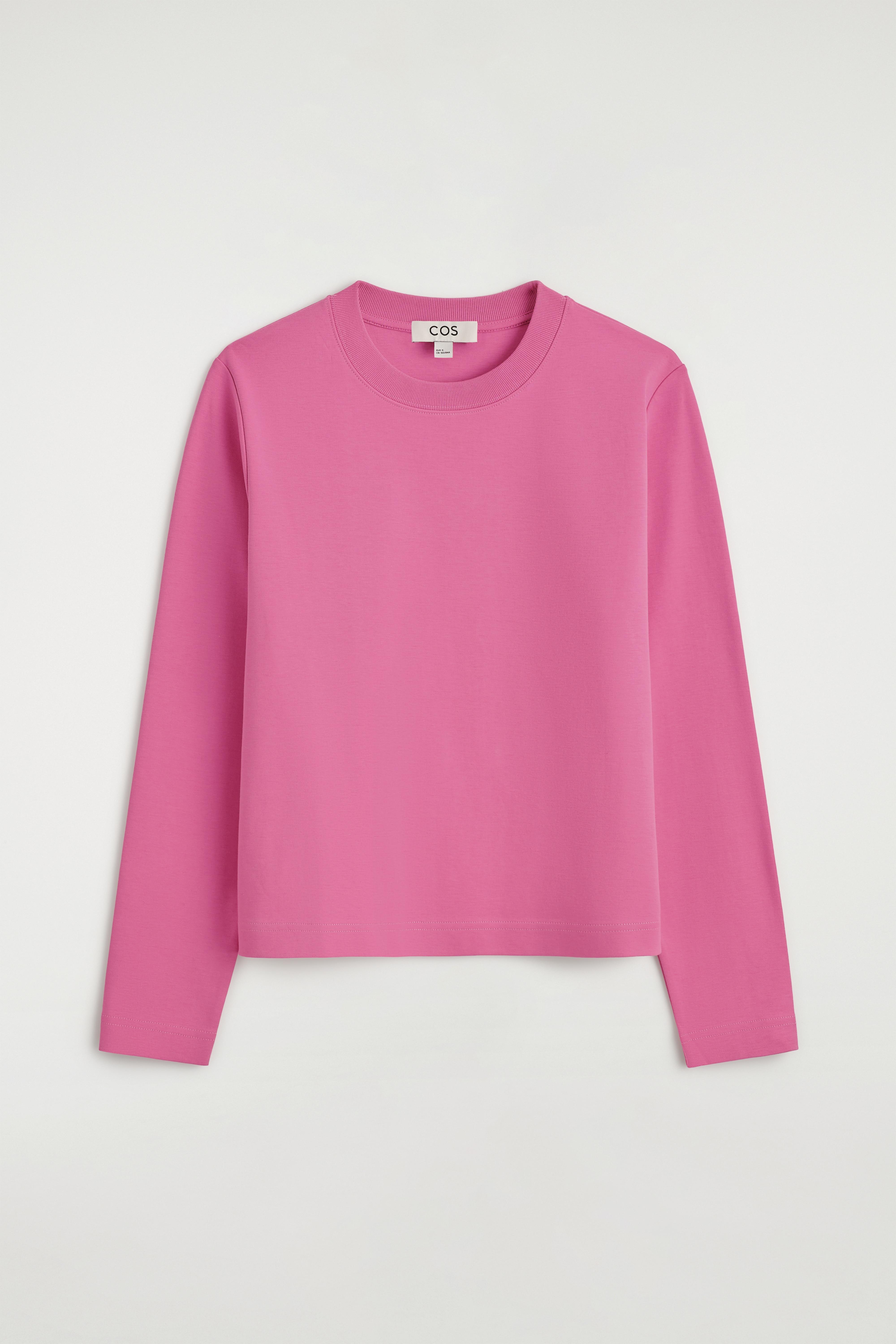 Un tricou roz cu mânecă lungă și guler rotund este purtat de model, asortat cu blugi negri și un pulover din tricot gri închis legat în jurul taliei.