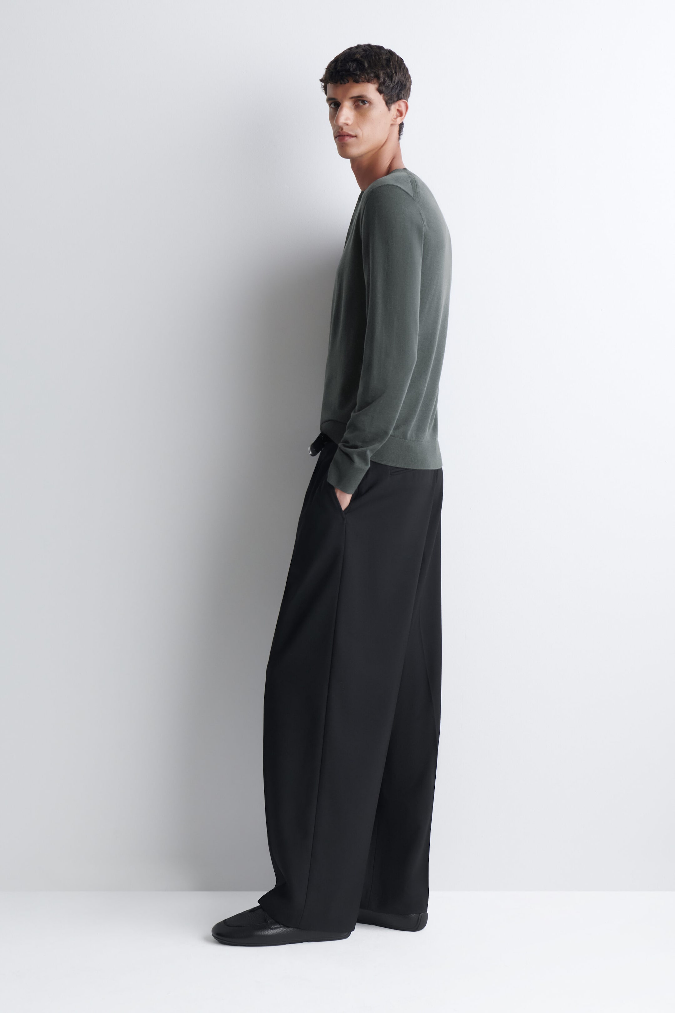A slim merino wool henley top in dark green is being worn by the model, who is standing in profile. The long-sleeved, fine-knit top is styled with wide-leg black trousers and black shoes.