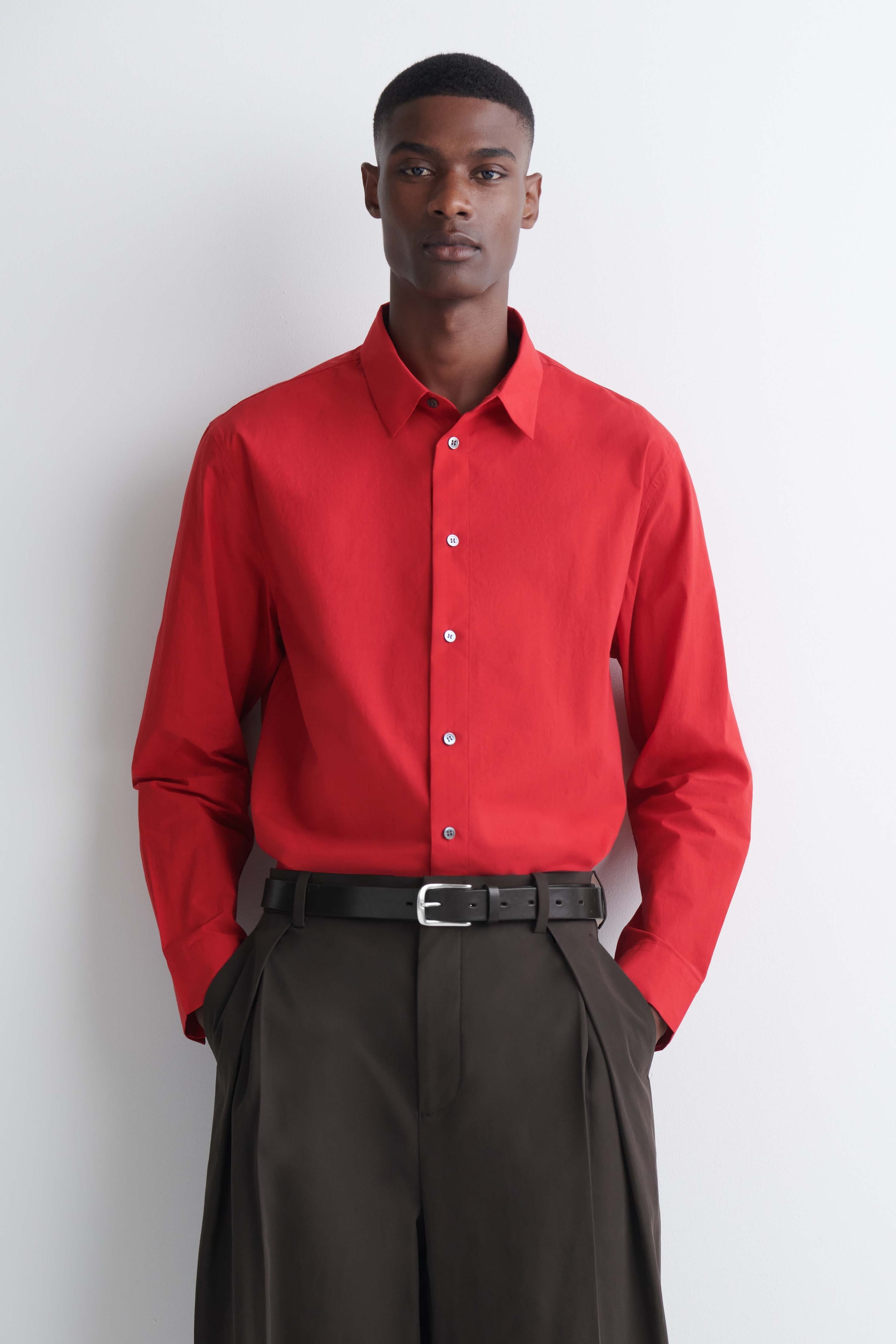 Ver imagen más grande: La modelo lleva una camisa roja de manga larga con cuello y cierre frontal de botones. La camisa está metida por dentro de unos pantalones plisados de color marrón oscuro, combinados con un cinturón de piel negro.