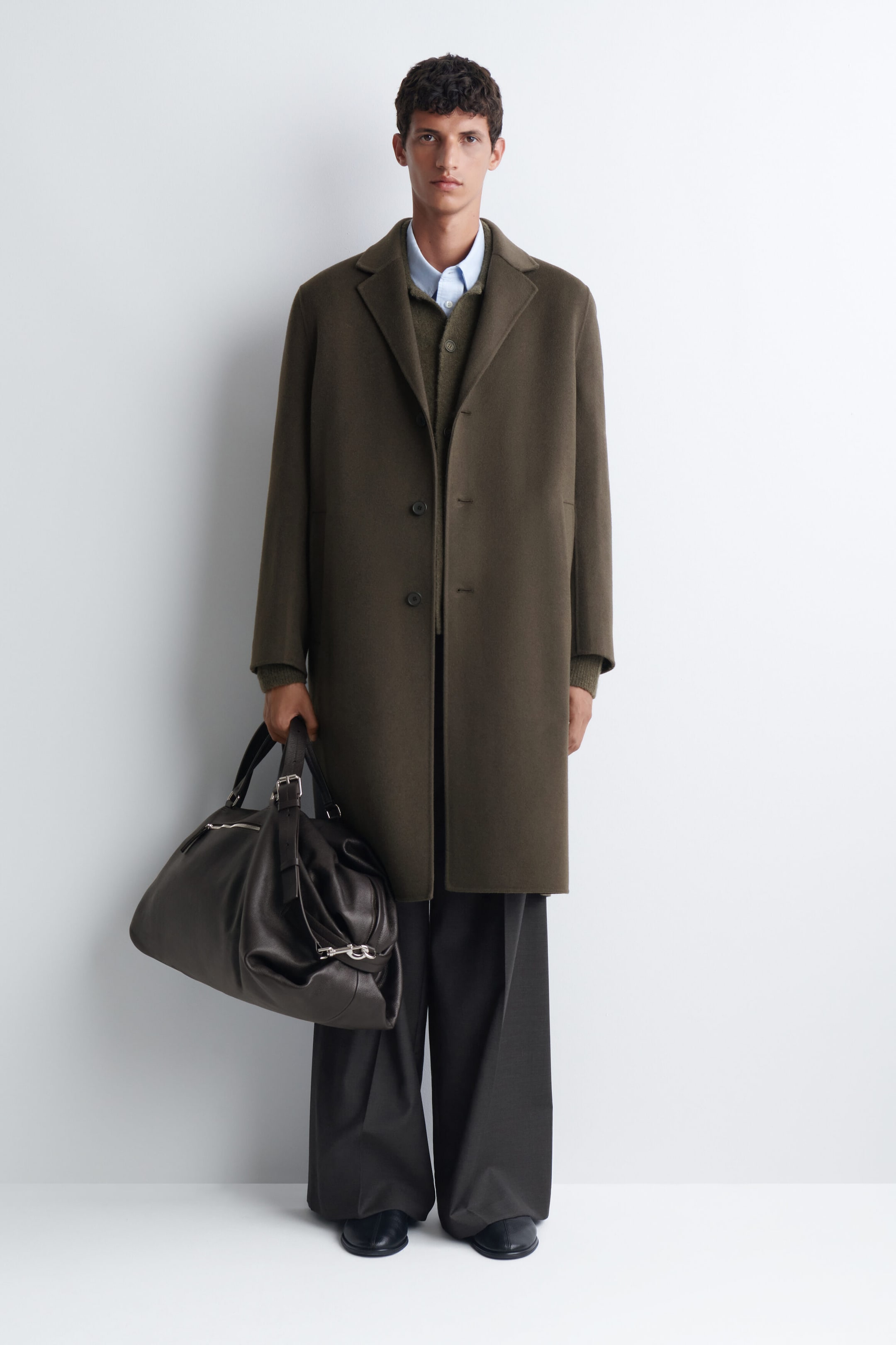 The Departure Duffle Bag in dark leather is being held by the model by its top handle. The bag features a slouchy, unstructured silhouette with dual handles and silver-toned metal hardware.