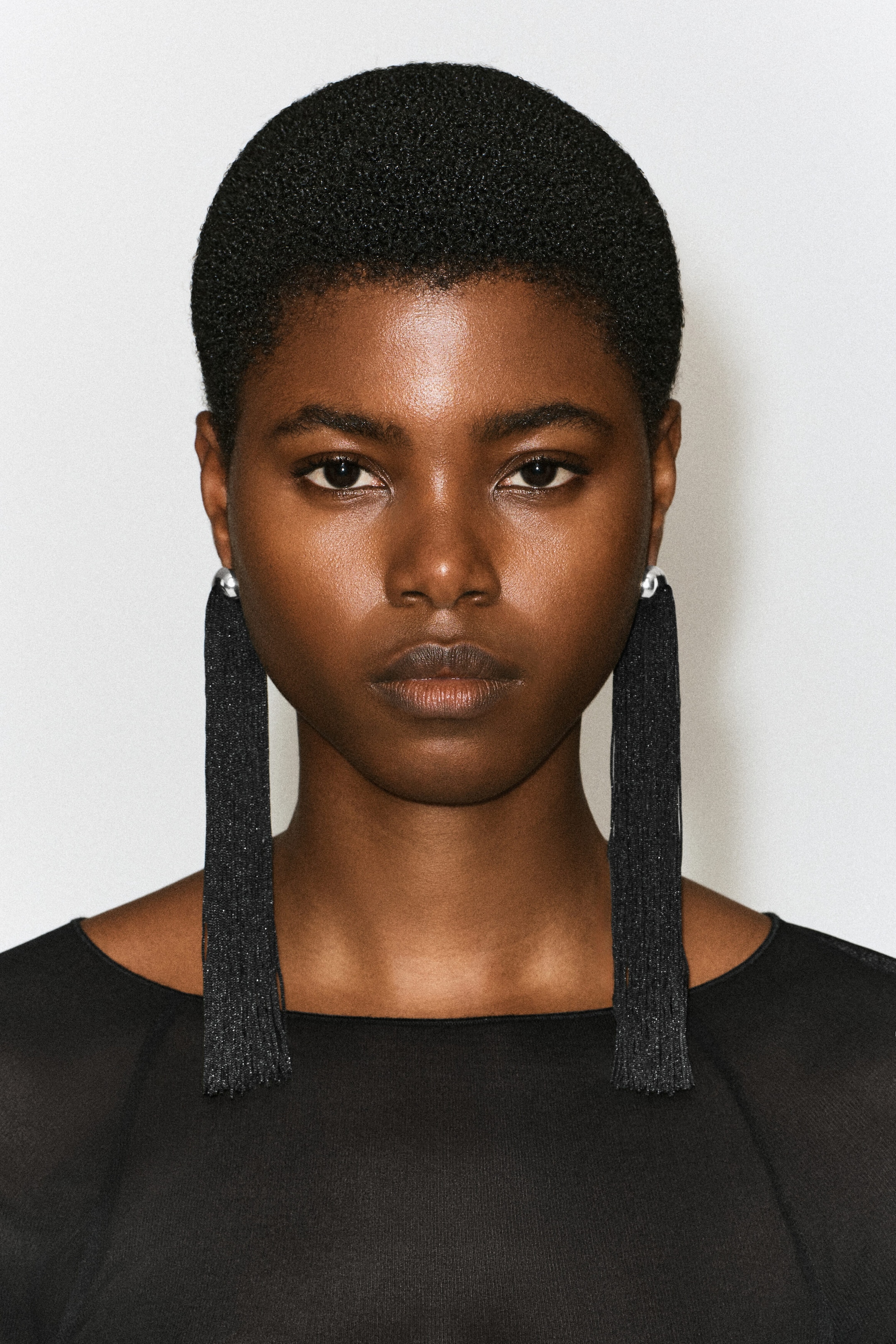 Fringed earrings featuring long, straight black tassels suspended from small, silver-toned dome studs are being worn by the model. The shoulder-length tassels are draping down over a black top.