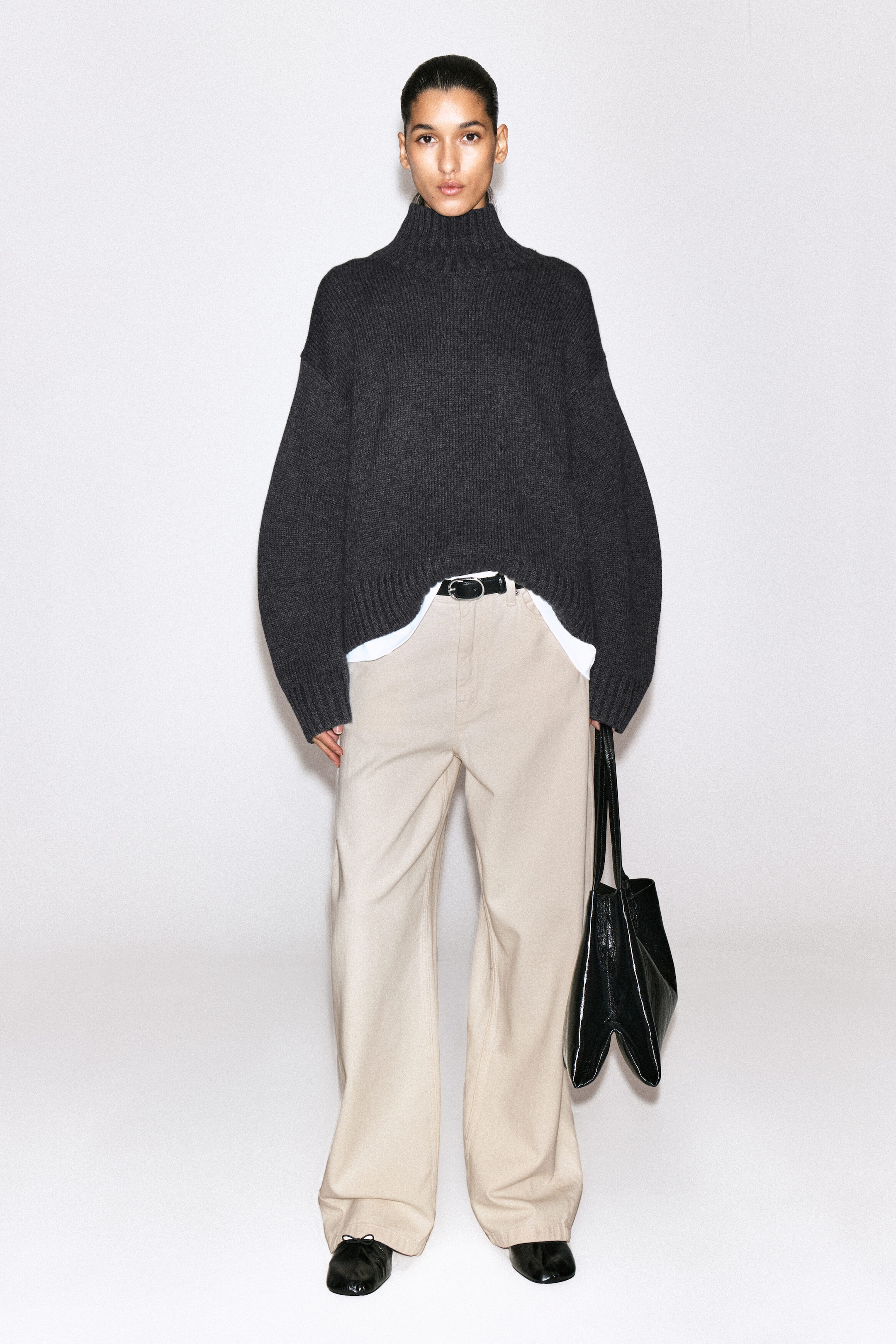 The Lagoon Wide-Leg Jeans are shown from the front, featuring a relaxed, wide-leg silhouette in light beige denim. The jeans sit at the waist with a black leather belt, which is partially visible beneath a dark grey roll-neck sweater tucked in at the front. The Lagoon Wide-Leg Jeans are shown from the front, featuring a relaxed, wide-leg silhouette in light beige denim. The jeans sit at the waist with a black leather belt, which is partially visible beneath a dark grey roll-neck sweater tucked in at the front.