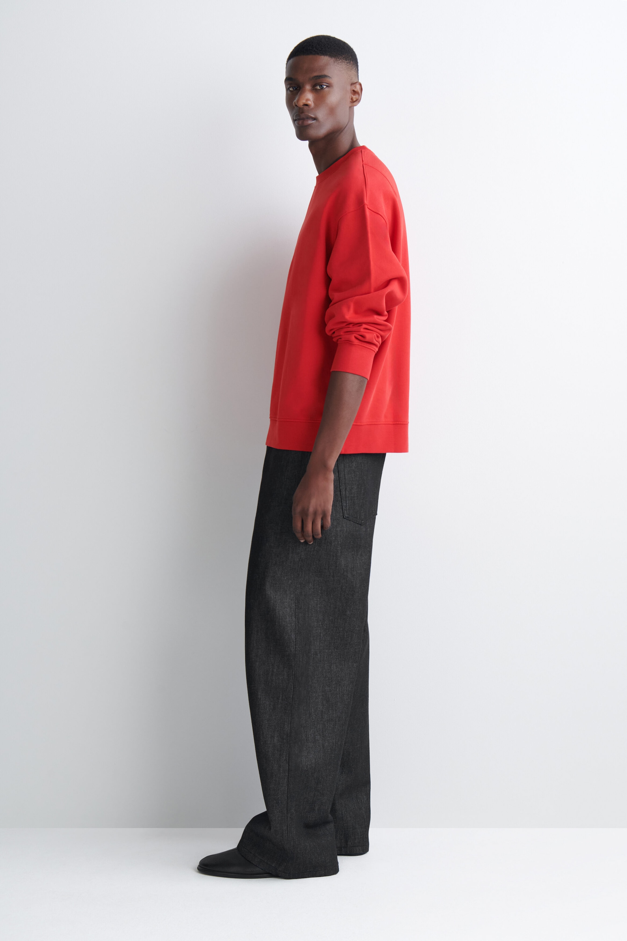 A red crew-neck sweatshirt with ribbed cuffs and hem is being worn by the model, who is standing in profile. The relaxed-fit sweatshirt is styled with dark-grey wide-leg trousers. A red crew-neck sweatshirt with ribbed cuffs and hem is being worn by the model, who is standing in profile. The relaxed-fit sweatshirt is styled with dark-grey wide-leg trousers.