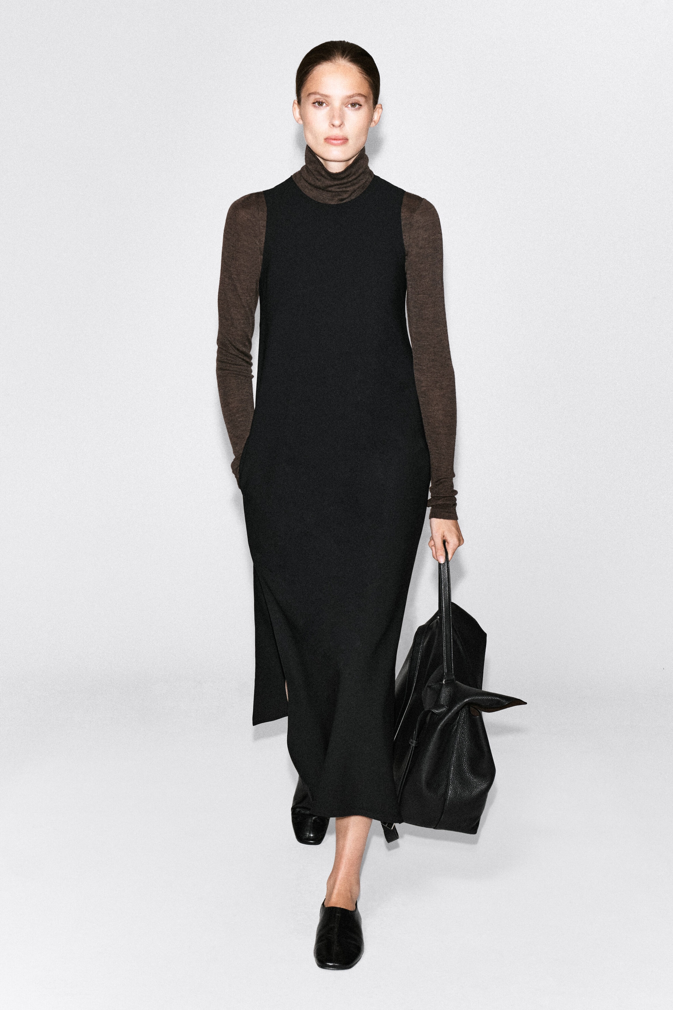 A sleeveless black crepe midi dress is worn over a brown long-sleeve turtleneck top. The dress features a straight silhouette, a high round neckline, and a side slit, and is styled with black flats and a black leather tote bag held by the model.