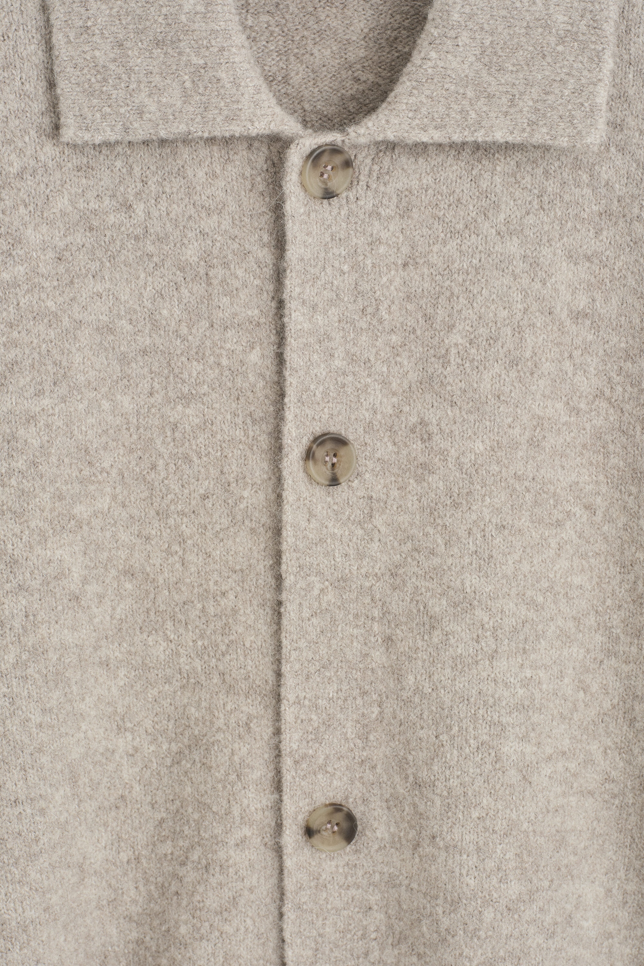 A close-up front view of a beige melange knit cardigan showing its soft, brushed texture and classic collar. The front placket features three visible mottled brown buttons. A close-up front view of a beige melange knit cardigan showing its soft, brushed texture and classic collar. The front placket features three visible mottled brown buttons.