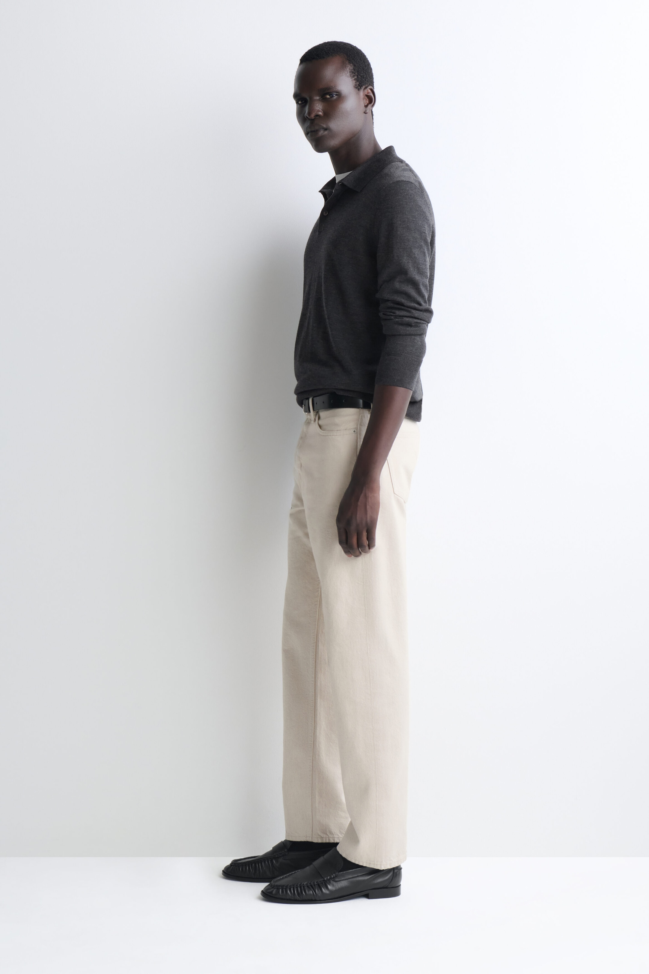 Das Modell steht nach links gewandt und trägt einen langärmeligen Strick-Polo-Pullover in Anthrazit mit klassischem Kragen und Knopfleiste. Der Pullover ist in eine cremefarbene, gerade geschnittene Hose gesteckt, die mit einem schwarzen Ledergürtel akzentuiert und mit schwarzen Leder-Loafers kombiniert ist. Das Modell steht nach links gewandt und trägt einen langärmeligen Strick-Polo-Pullover in Anthrazit mit klassischem Kragen und Knopfleiste. Der Pullover ist in eine cremefarbene, gerade geschnittene Hose gesteckt, die mit einem schwarzen Ledergürtel akzentuiert und mit schwarzen Leder-Loafers kombiniert ist.